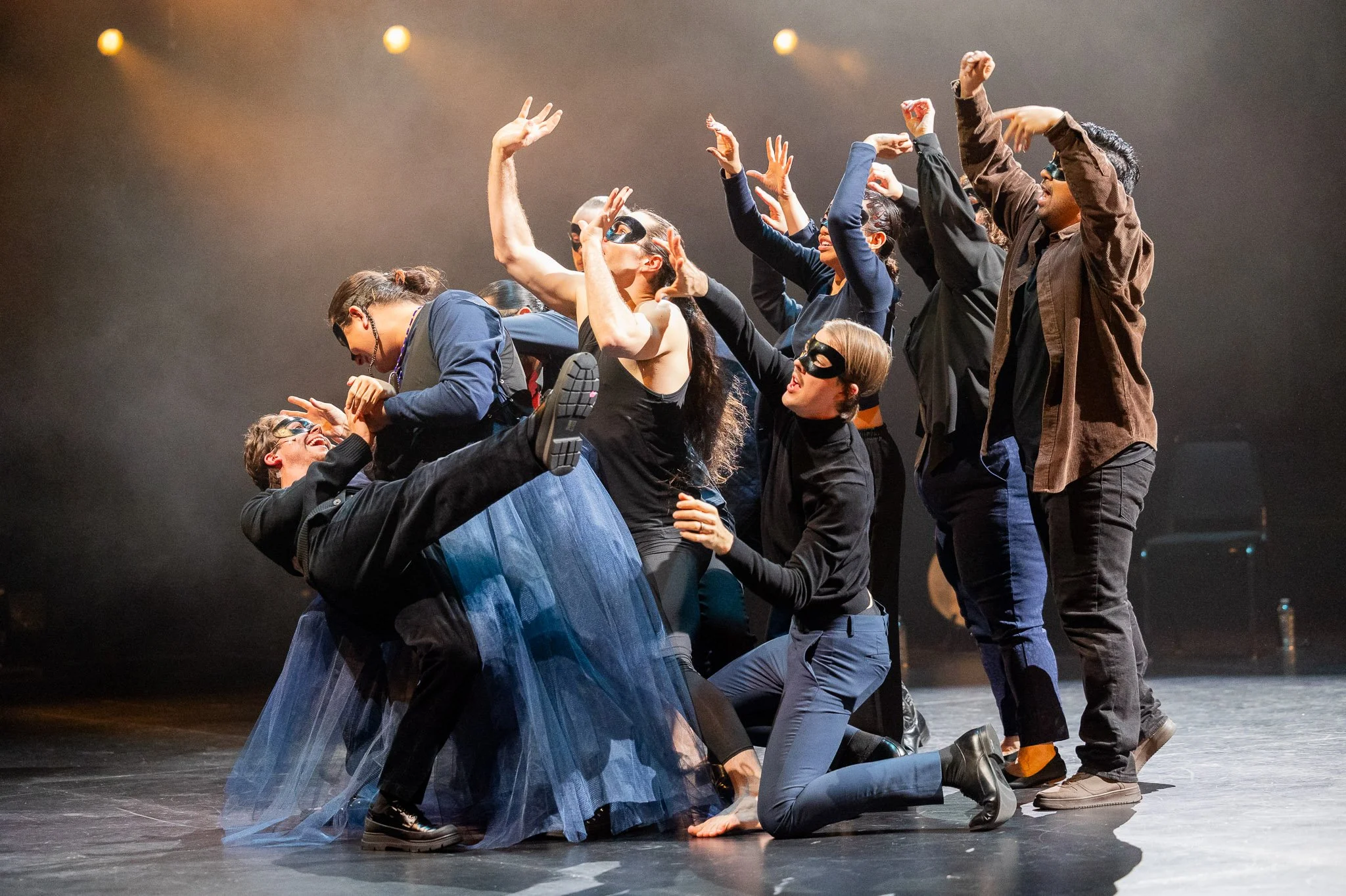 In the centre of a cluster of six people, a masculine figure dressed in a long blue tulle skirt crouches. They caress the face of another man. The surrounding figures raise their hands to the ceiling in supplication or dance. 