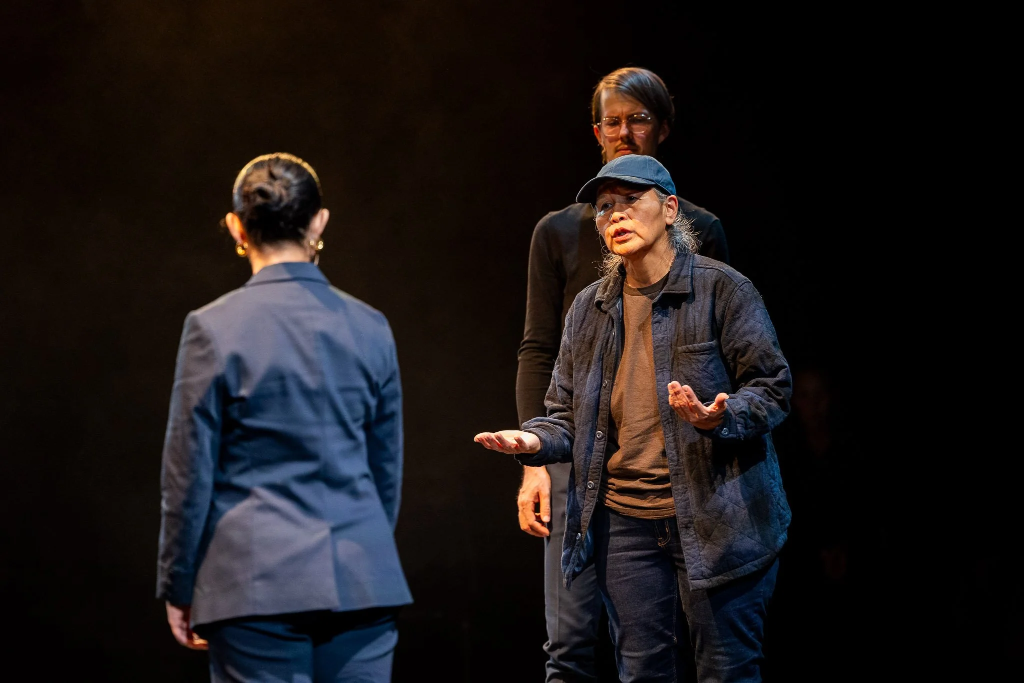 A woman in a blue plaid coat and brown t-shirt converses with another woman in a navy suit. In the background, a masculine figure in a black sweater observes the conversation. 