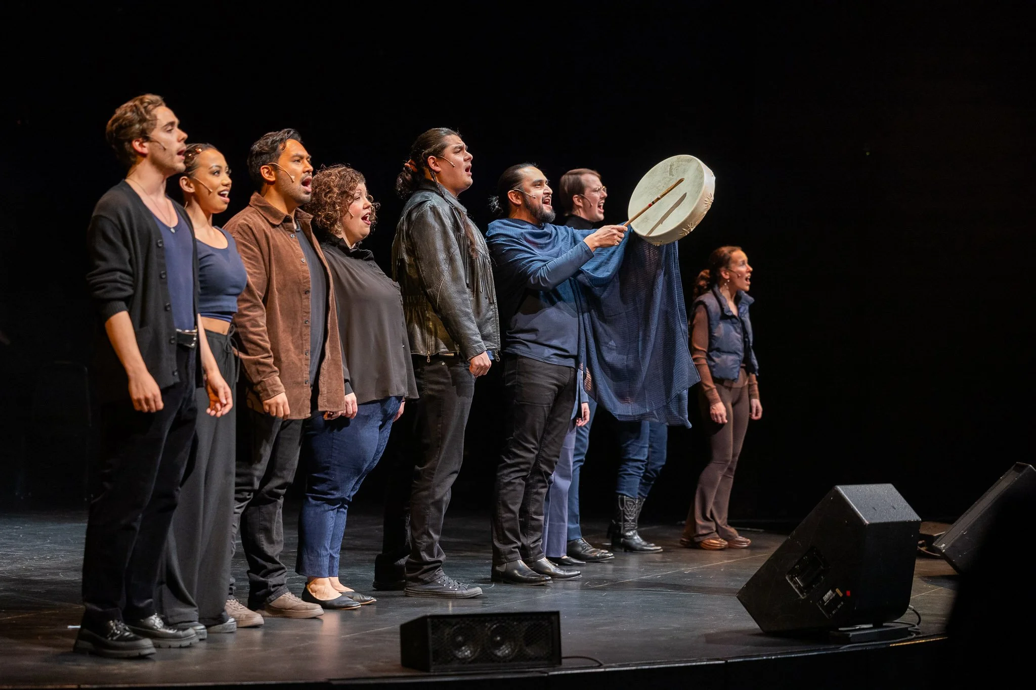 Nine performers stand in a line at the foot of the stage wearing shades of navy, brown, and black. One of them holds out a leather drum. All are singing. 