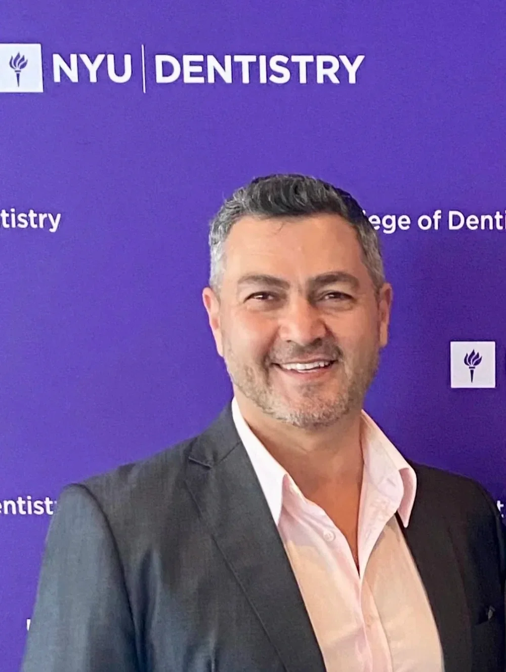 A man with short, salt-and-pepper hair wearing a suit jacket and white shirt, standing in front of a purple NYU Dentistry backdrop.