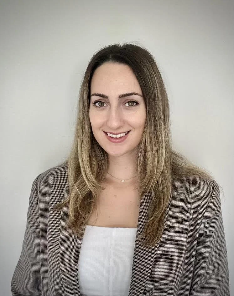 A woman with long, wavy blonde hair wearing a brown plaid blazer and a white top, smiling against a plain light-colored background.
