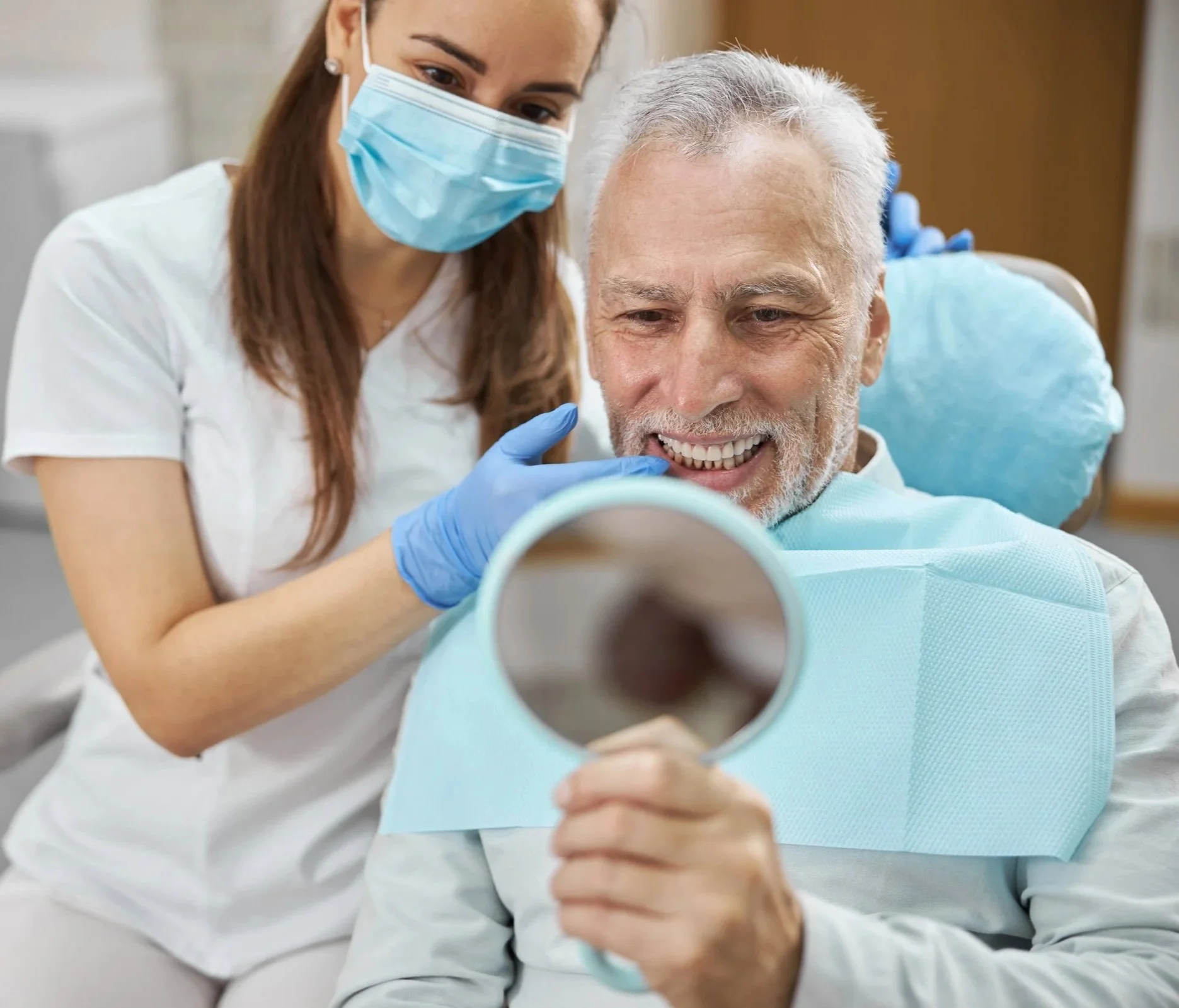 A female healthcare worker wearing a face mask and gloves is assisting an elderly man in a dental or medical setting, who is smiling and holding a mirror as he examines his teeth.