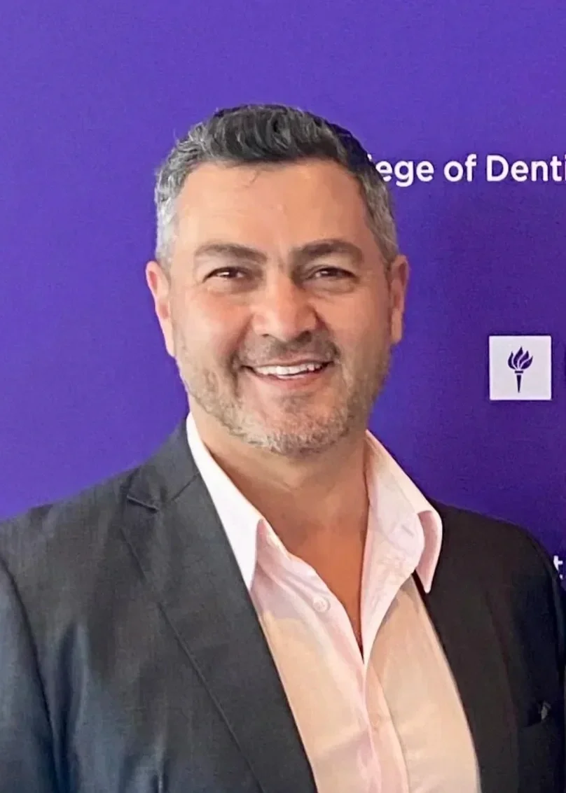 A smiling man in a suit with a pink shirt standing in front of a purple background with white text and symbols.
