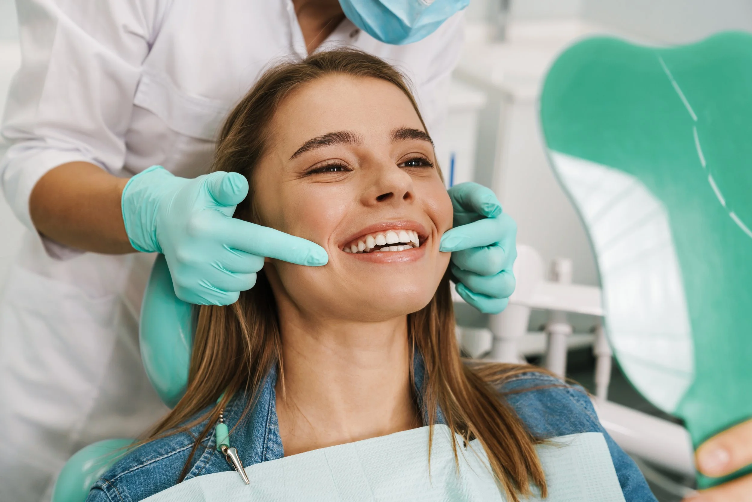 A woman at the dentist smiling as a dentist inspects her teeth, with her holding a mirror and wearing a dental bib.
