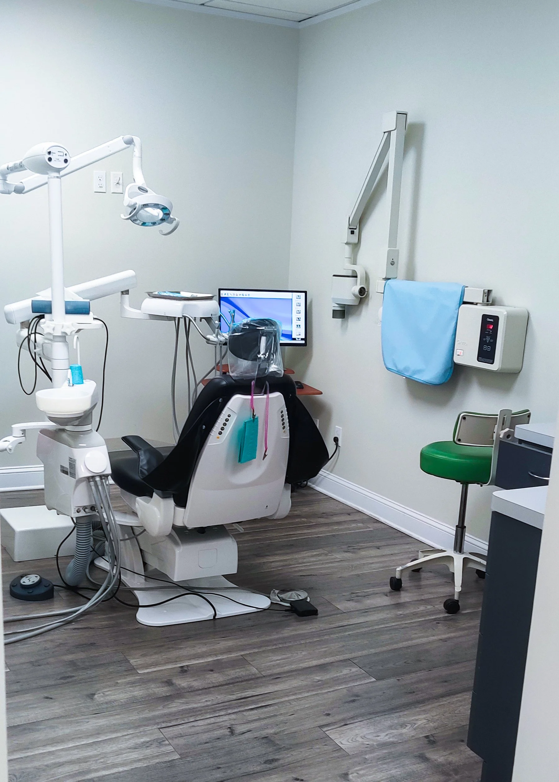 A modern dental examination room with dental chair, overhead light, monitor, medical equipment, and green stool.