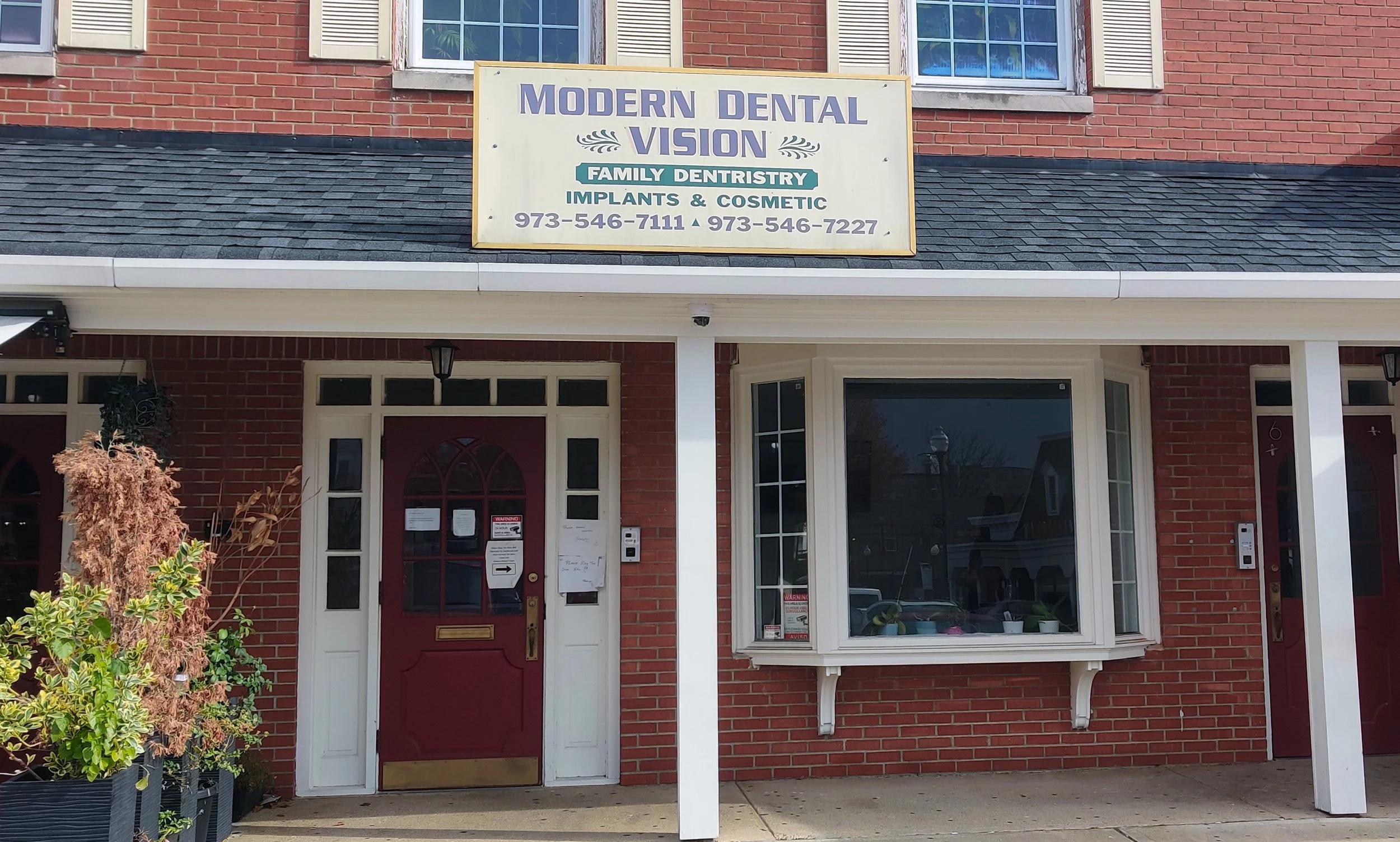 Exterior of a dental office with a yellow sign reading 'Modern Dental Vision' and contact numbers, brick building, red door, large window with plants inside.