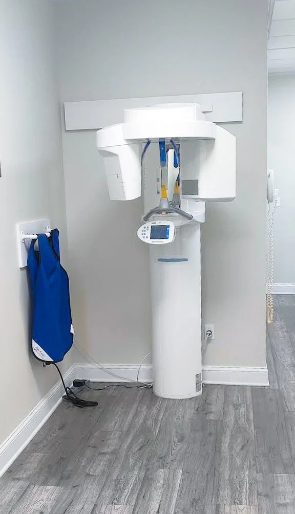 A dental X-ray machine in a medical clinic or hospital room with gray walls and gray wood flooring.