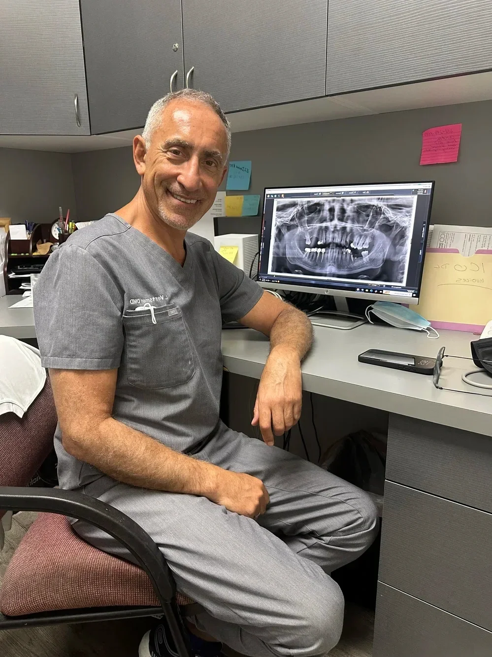 A smiling male dentist sitting in an office at a desk with a computer showing a dental X-ray of a patient's teeth.