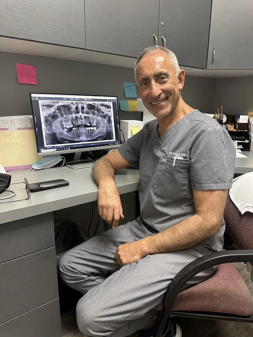 A smiling male dentist in gray scrubs sitting at a desk in front of a computer monitor displaying a dental X-ray of a patient's teeth and jaw.