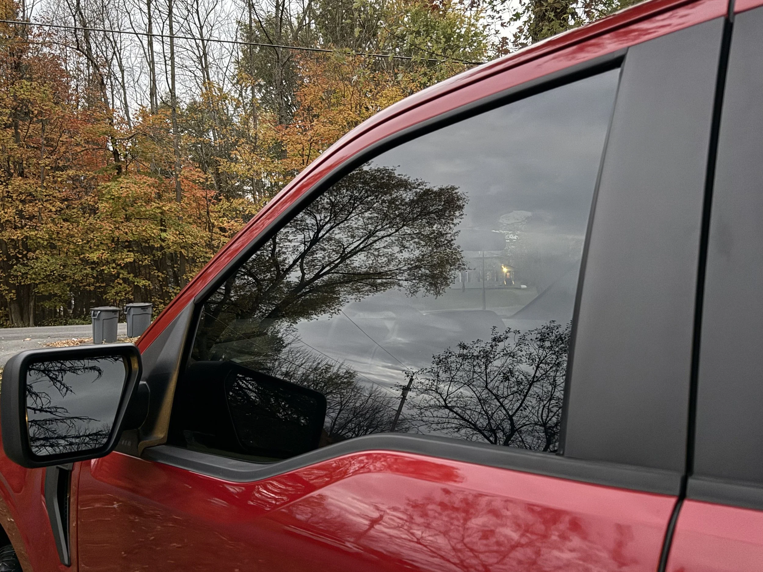 A close-up of a red vehicle's side, showing the side mirror and window reflection of trees with autumn-colored leaves and a cloudy sky.