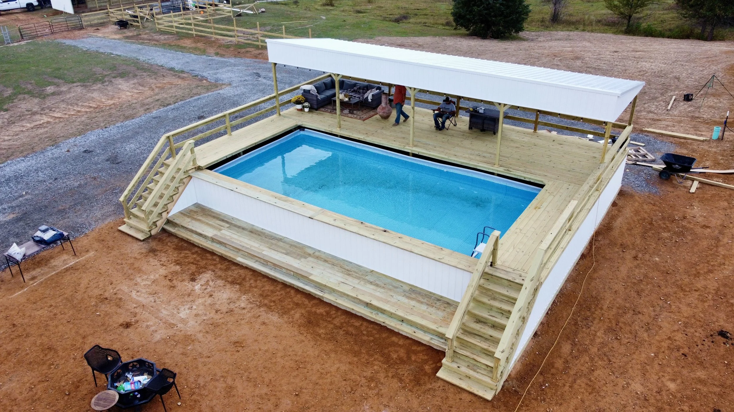 An outdoor swimming pool surrounded by a wooden deck with stairs, under construction. The deck features a shaded seating area with furniture, and the setting is a backyard with some bare soil and greenery in the background.