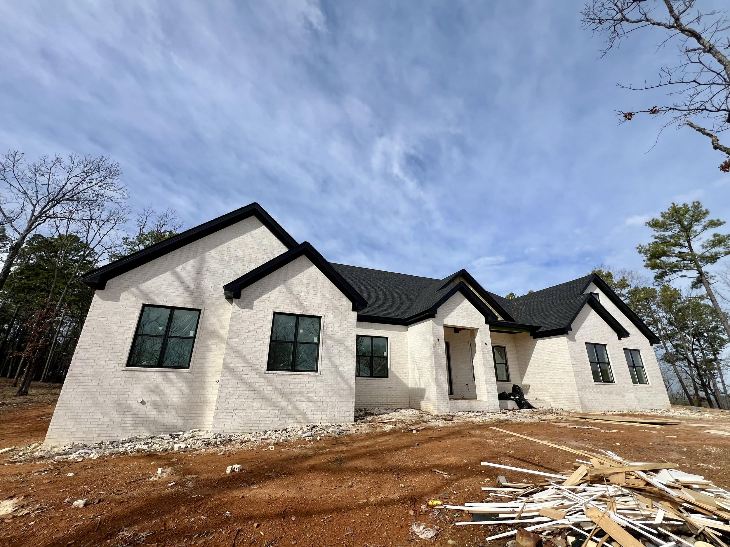 Newly constructed white brick house with black roof on a dirt lot, surrounded by trees, with construction materials in front.