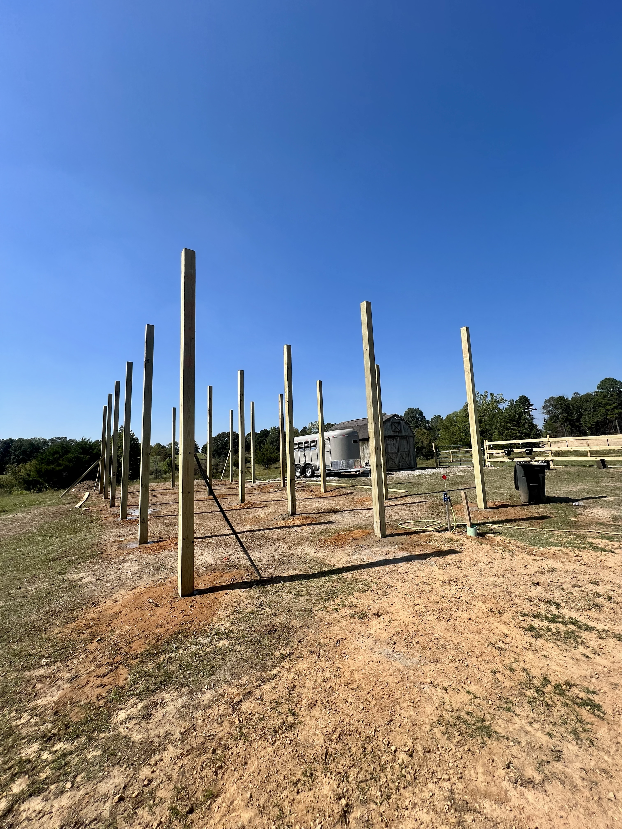Construction site with vertical wooden posts on dirt ground, under a clear blue sky, with a trailer, shed, and trees in the background.