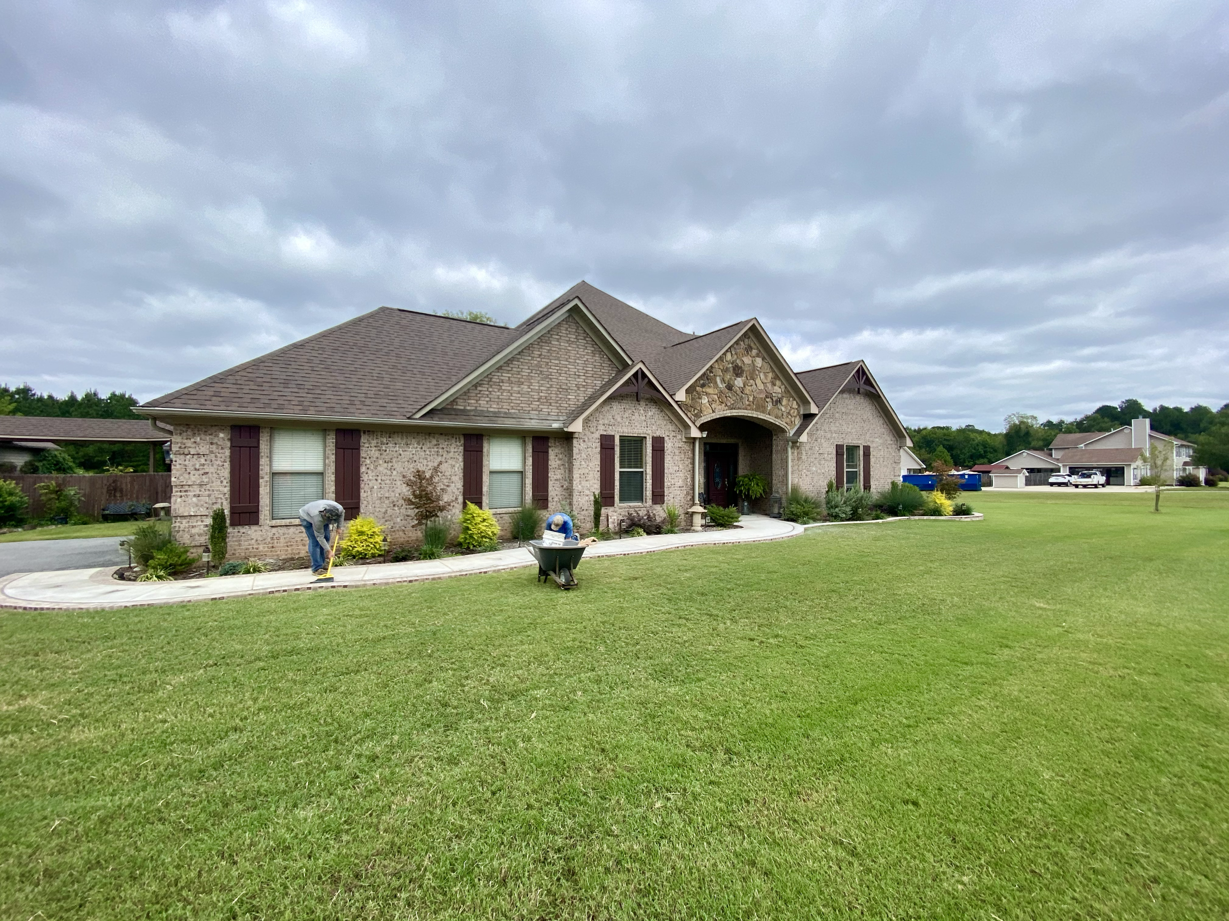 A modern brick house with a brown shingle roof, purple window shutters, and a landscaped front yard. Two workers are laying a concrete sidewalk, and there is greenery including small trees and bushes.