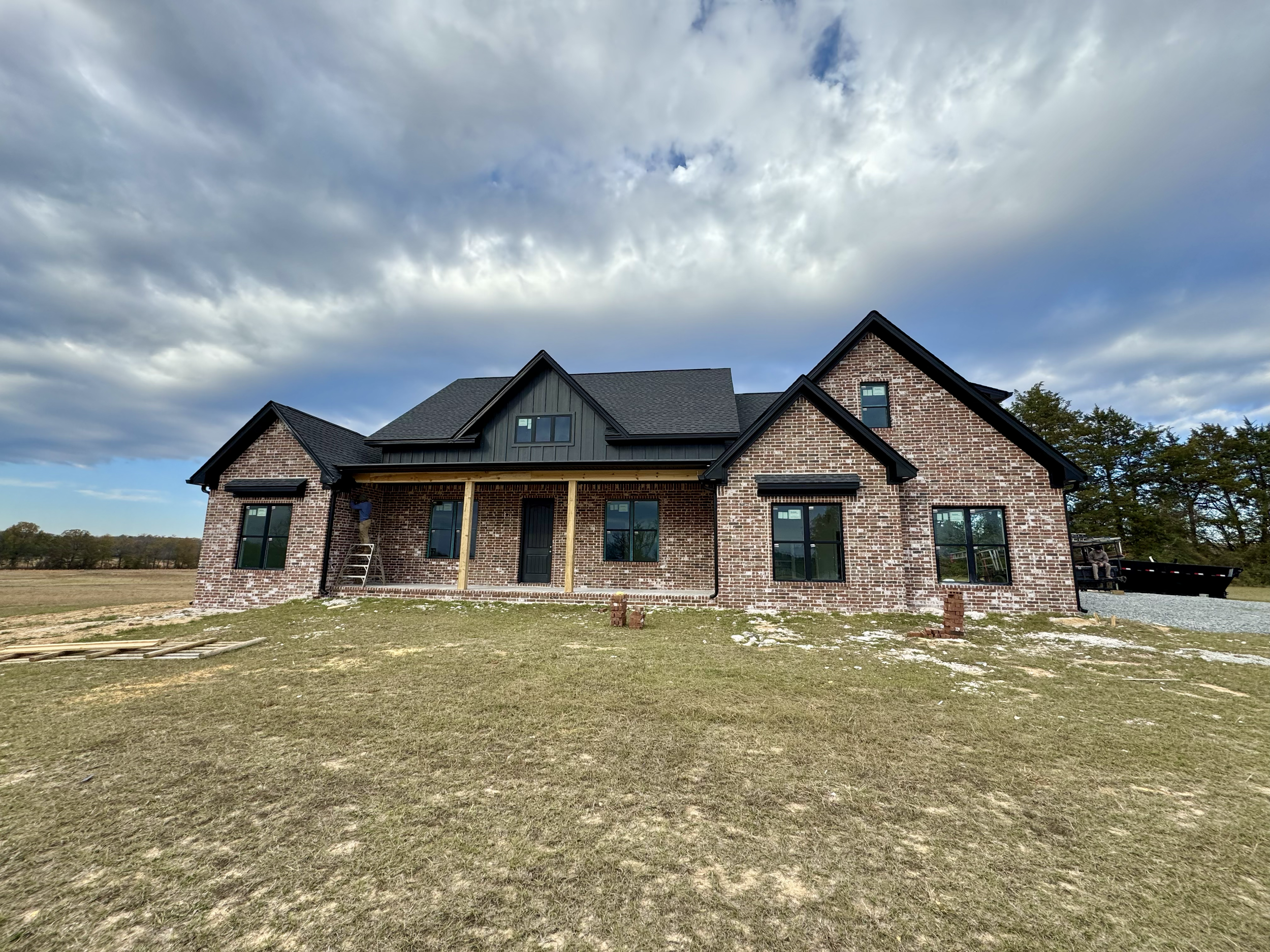 New brick house under construction with black windows and a black roof, located in a grassy field with a cloudy sky above.
