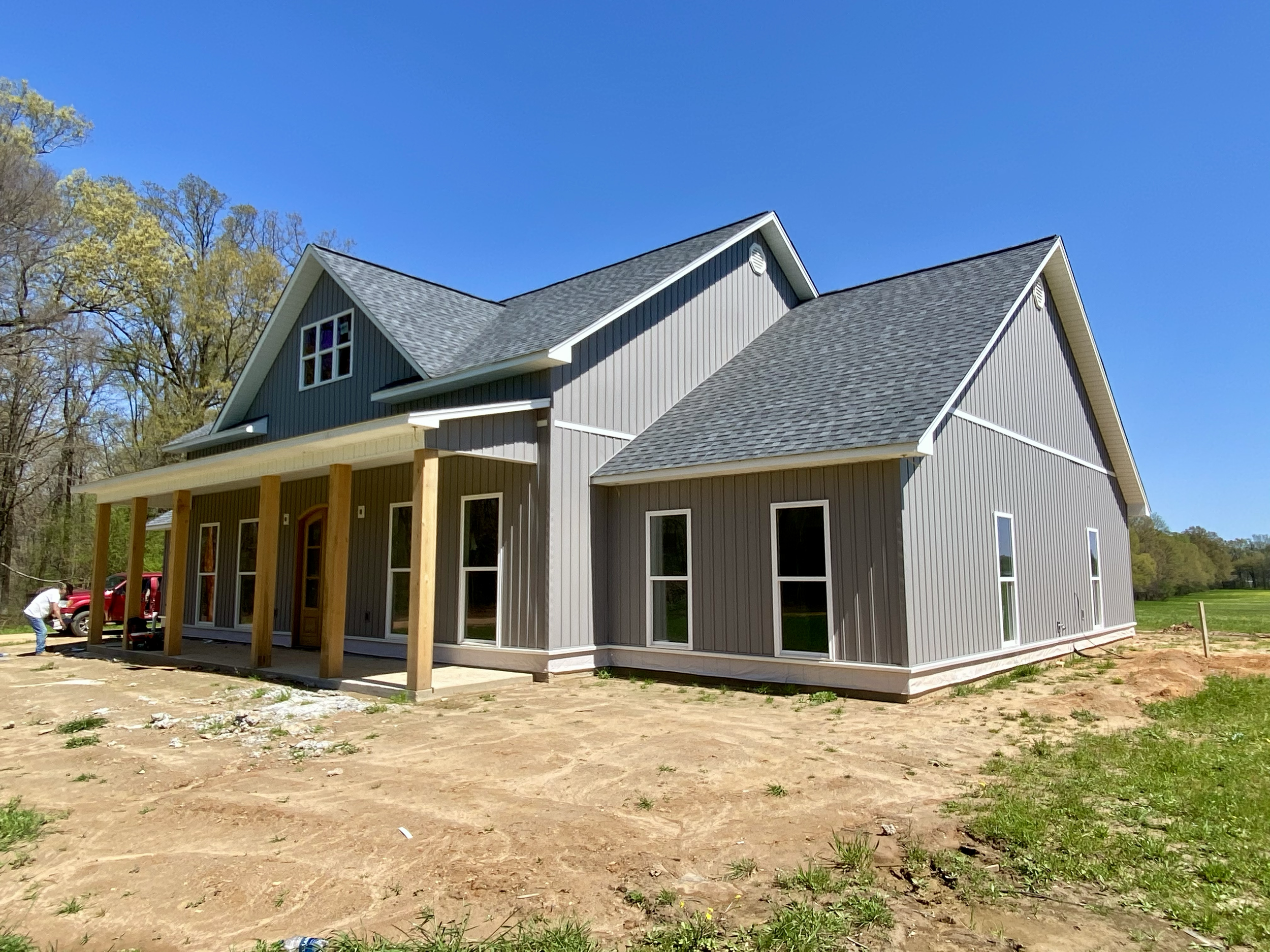 New gray house with large windows, a covered porch with wooden posts, and a dark shingled roof, surrounded by a dirt yard with some patches of grass, under a clear blue sky.