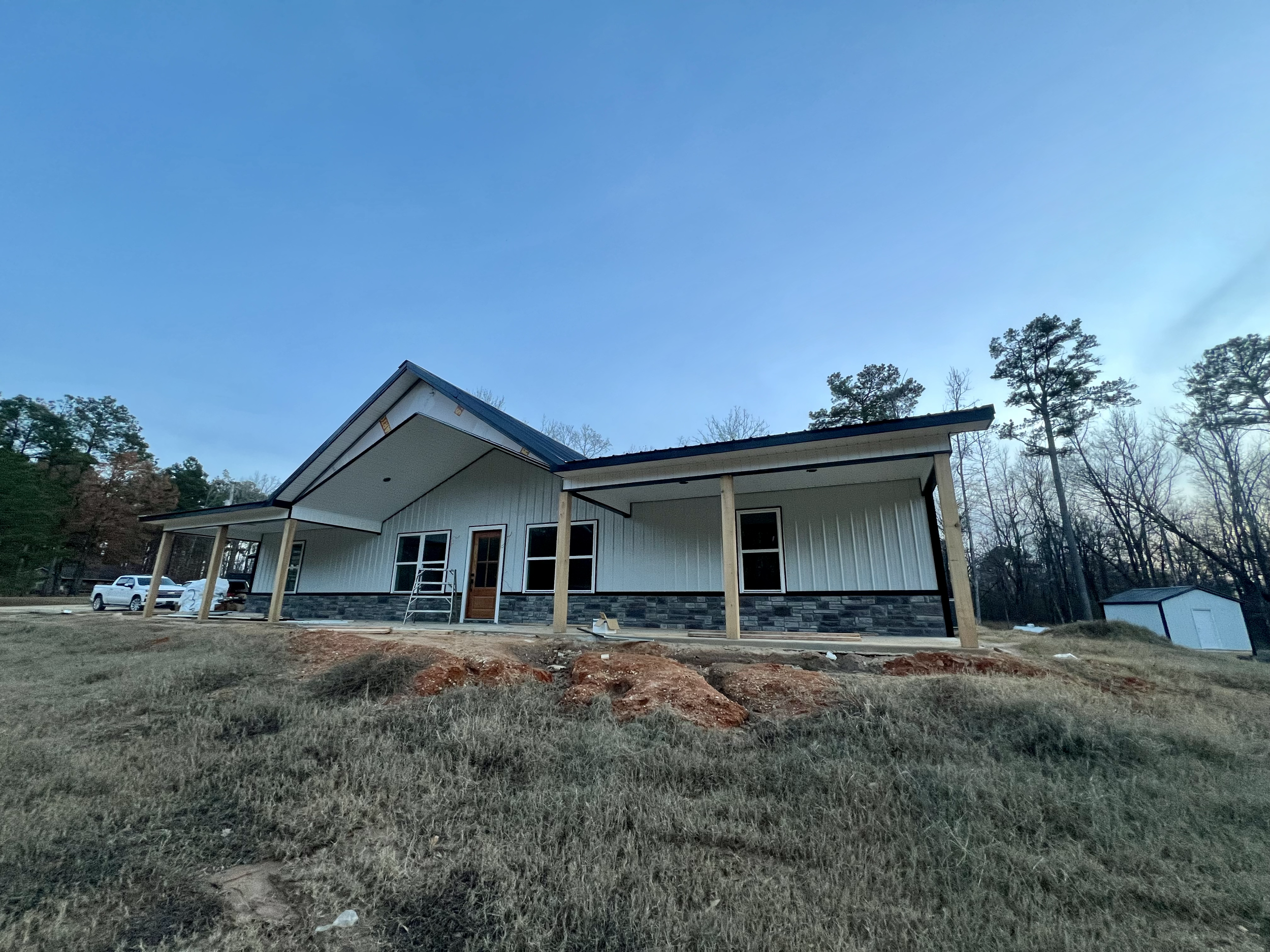 Newly constructed house with white siding, stone accents, and a metal roof, situated on a grassy plot with some dirt patches, surrounded by tall trees under a clear blue sky.