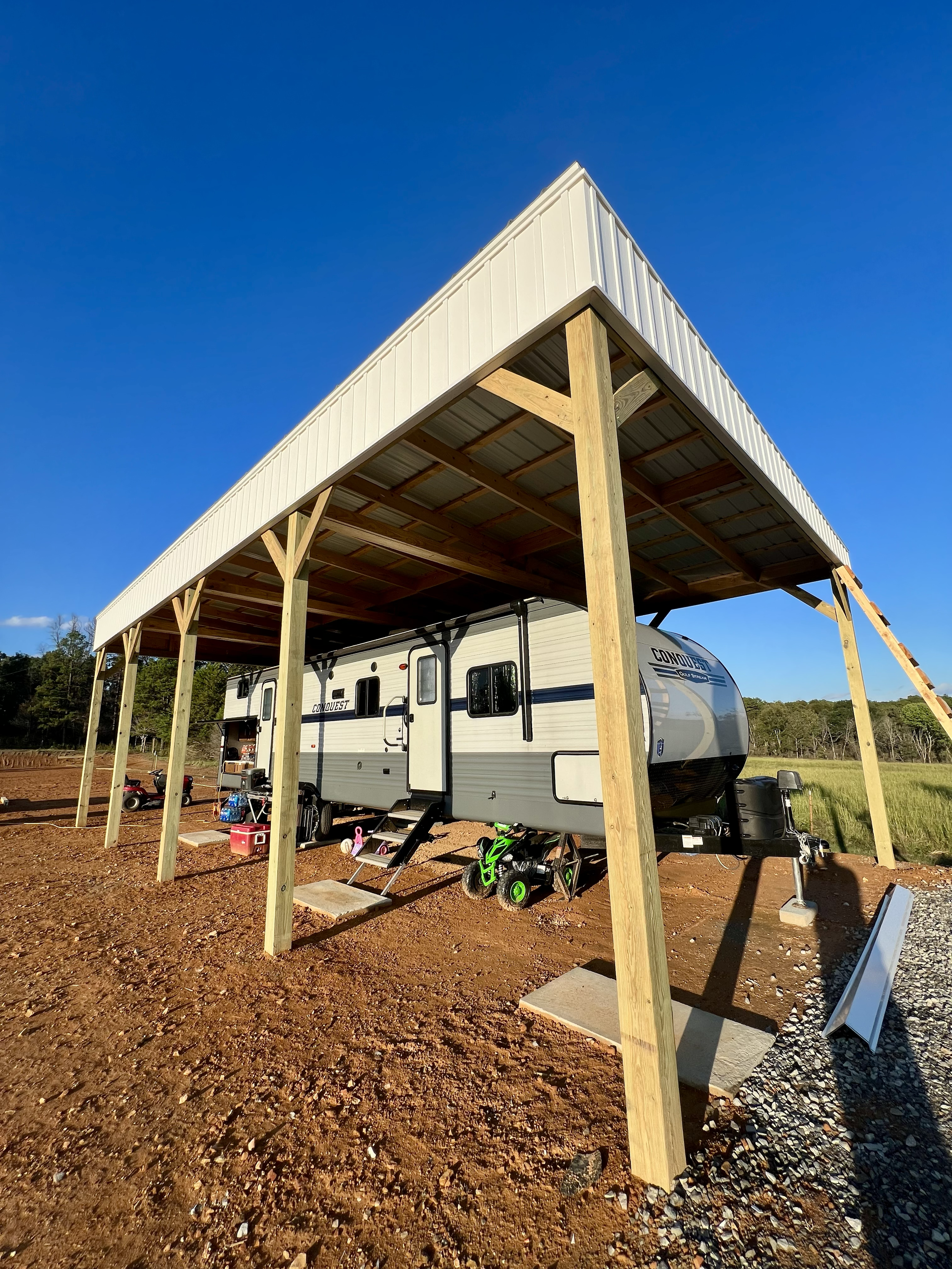 A travel trailer parked under a large wooden carport with a metal roof on a dirt lot, with a clear blue sky and green trees in the background.