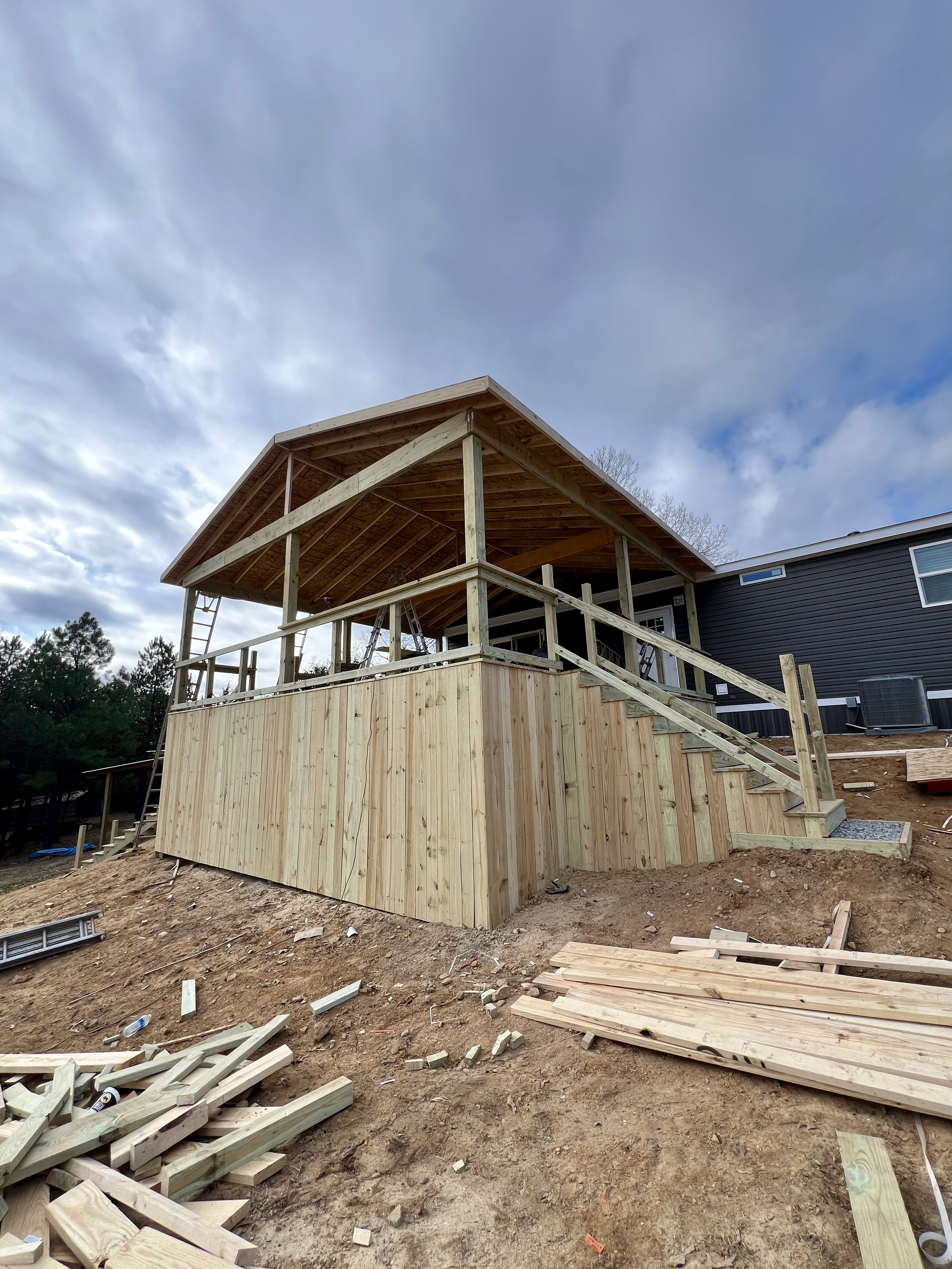 A house under construction with a wooden deck and railing in progress, surrounded by construction materials and tools on uneven dirt ground.
