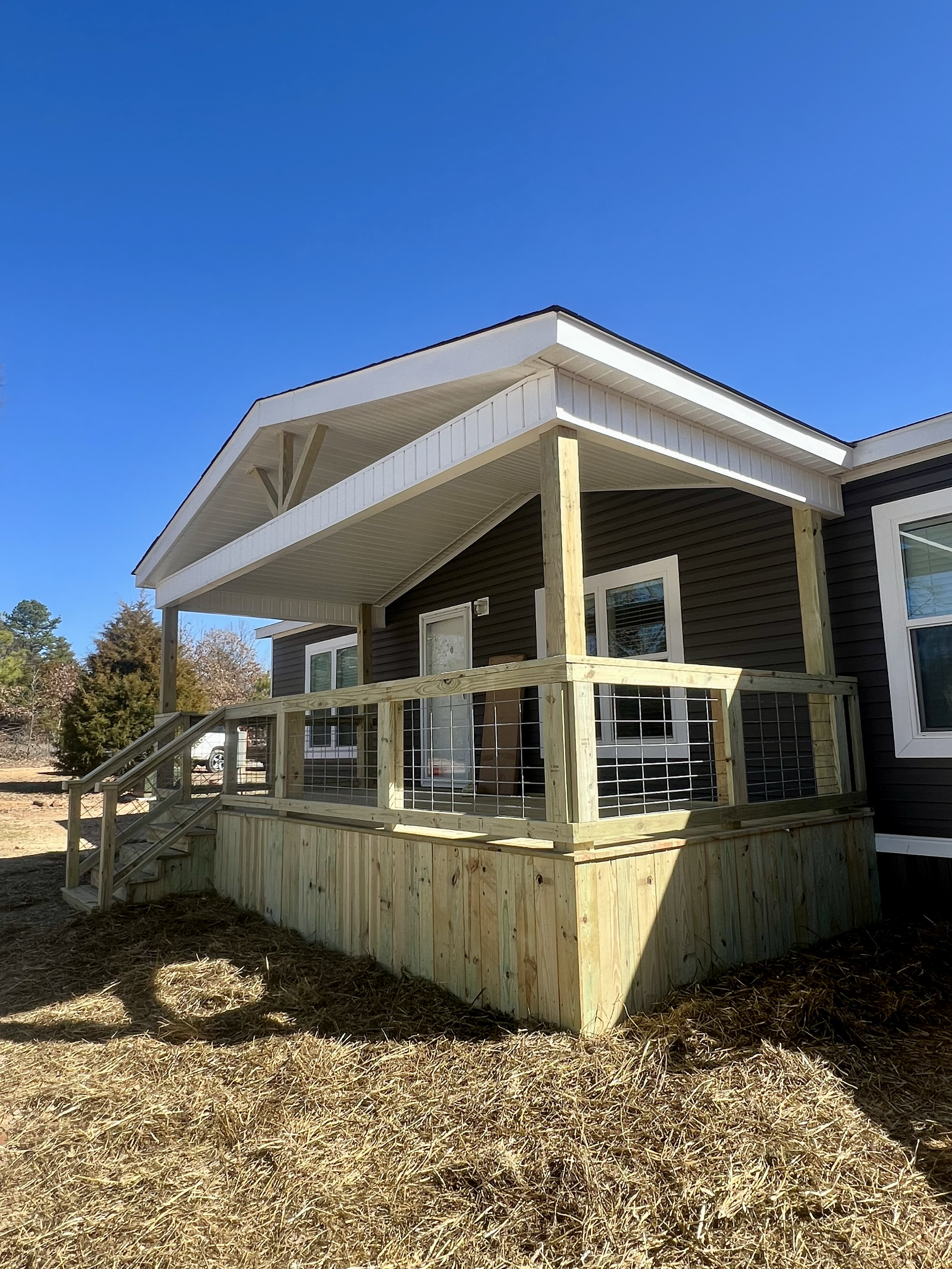 Newly constructed wooden deck attached to a house with dark siding, featuring a covered porch with railing and steps, under a clear blue sky.