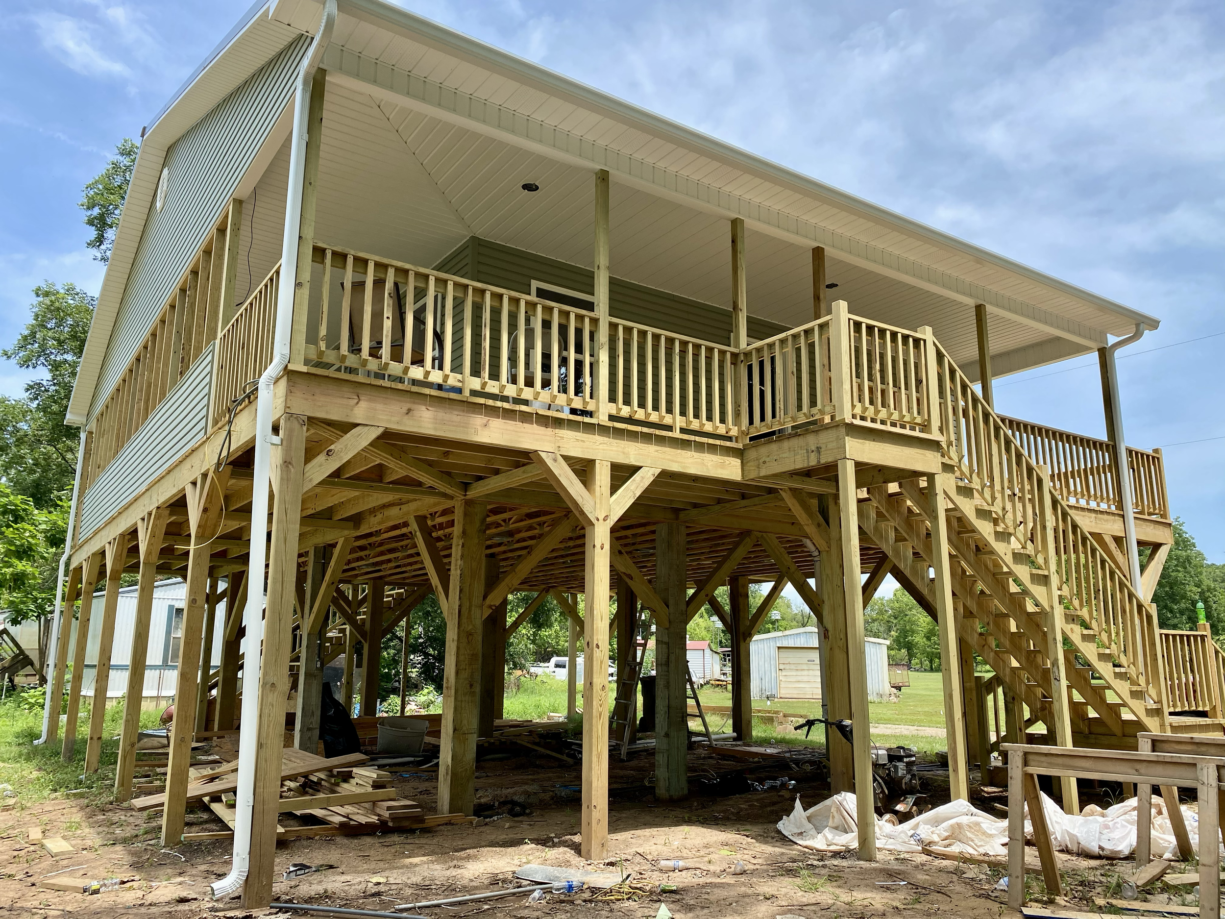 A two-story house under construction with a wooden deck and staircase, surrounded by construction materials and equipment, with a clear sky and trees in the background.