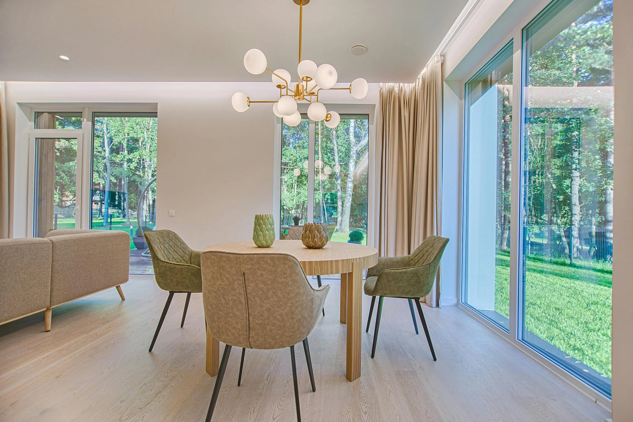 Modern dining area with round wooden table, four upholstered chairs, decorative vases, large windows with beige curtains, and a stylish chandelier, overlooking a lush green outdoor space.