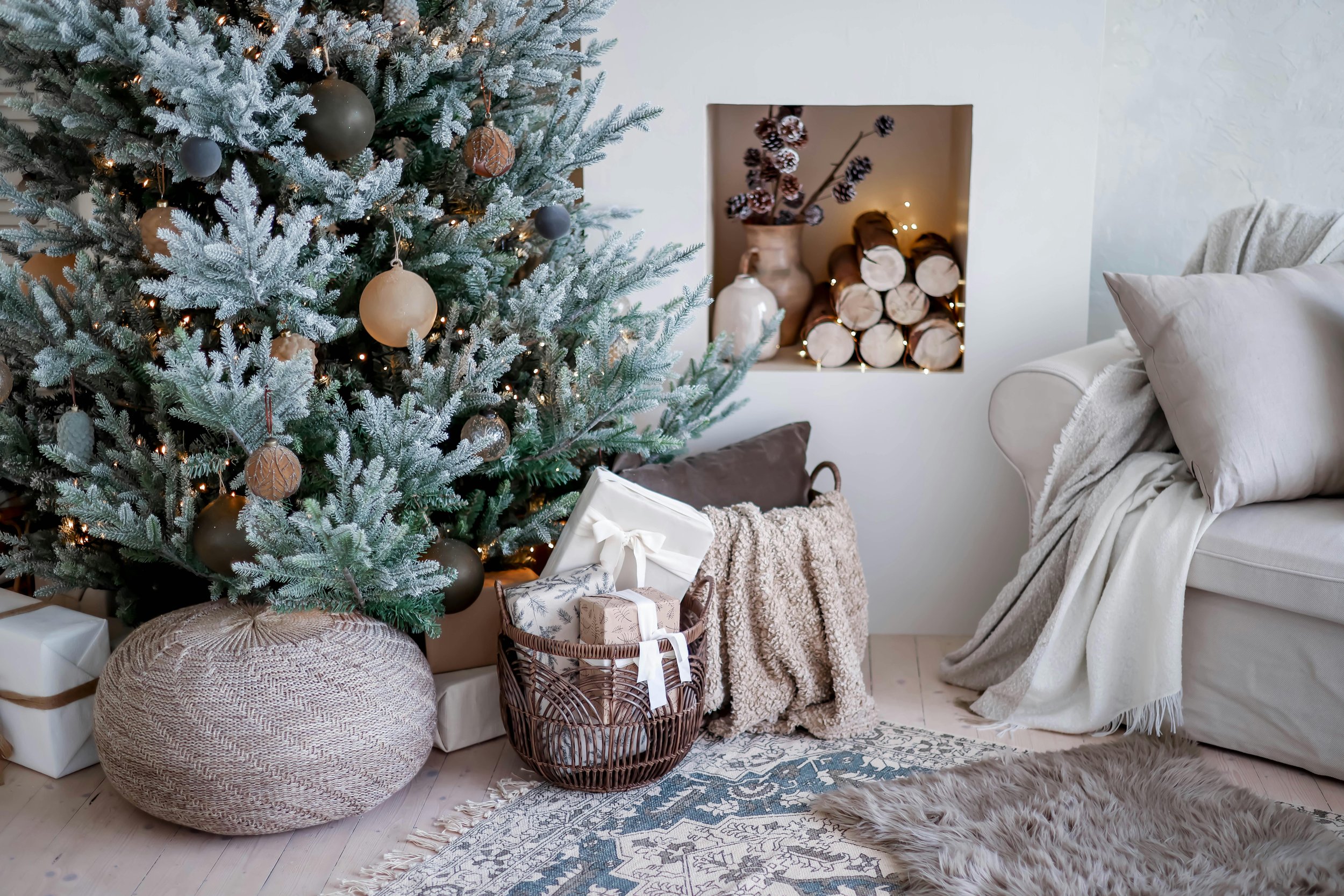 Decorated Christmas tree with ornaments and lights, gift boxes underneath, cozy living room corner with a couch, throw blanket, rug, and a niche with firewood and decorative items.