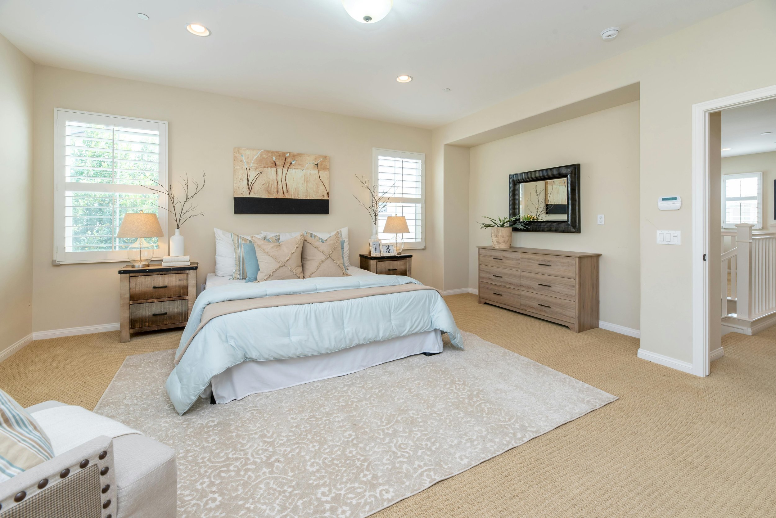 A bright bedroom with beige walls, a white bed with beige and blue pillows, two side tables with lamps, a beige rug, a wooden dresser with a mirror, and windows with white shutters.
