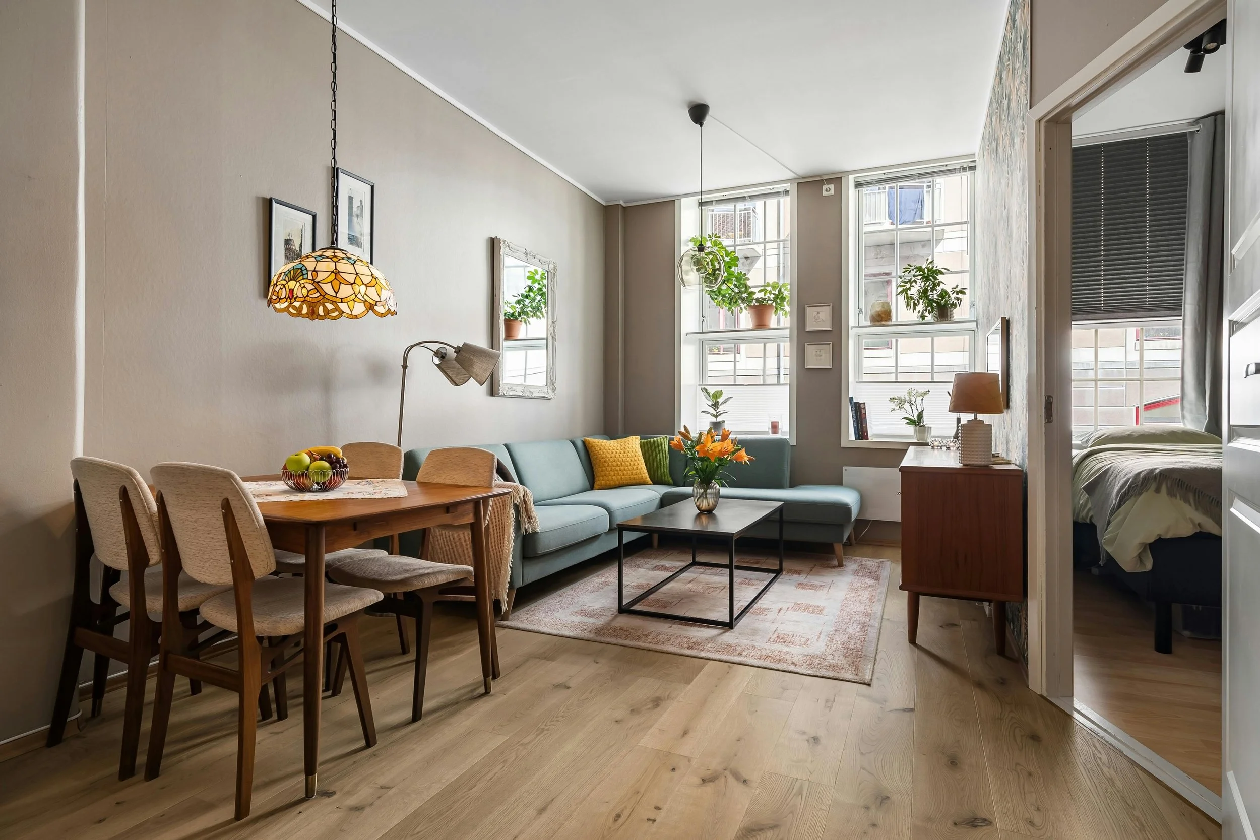 Living room with a light blue sectional sofa, wooden dining table with four chairs, and large windows with potted plants. A doorway leads to a bedroom with a bed and blanket.