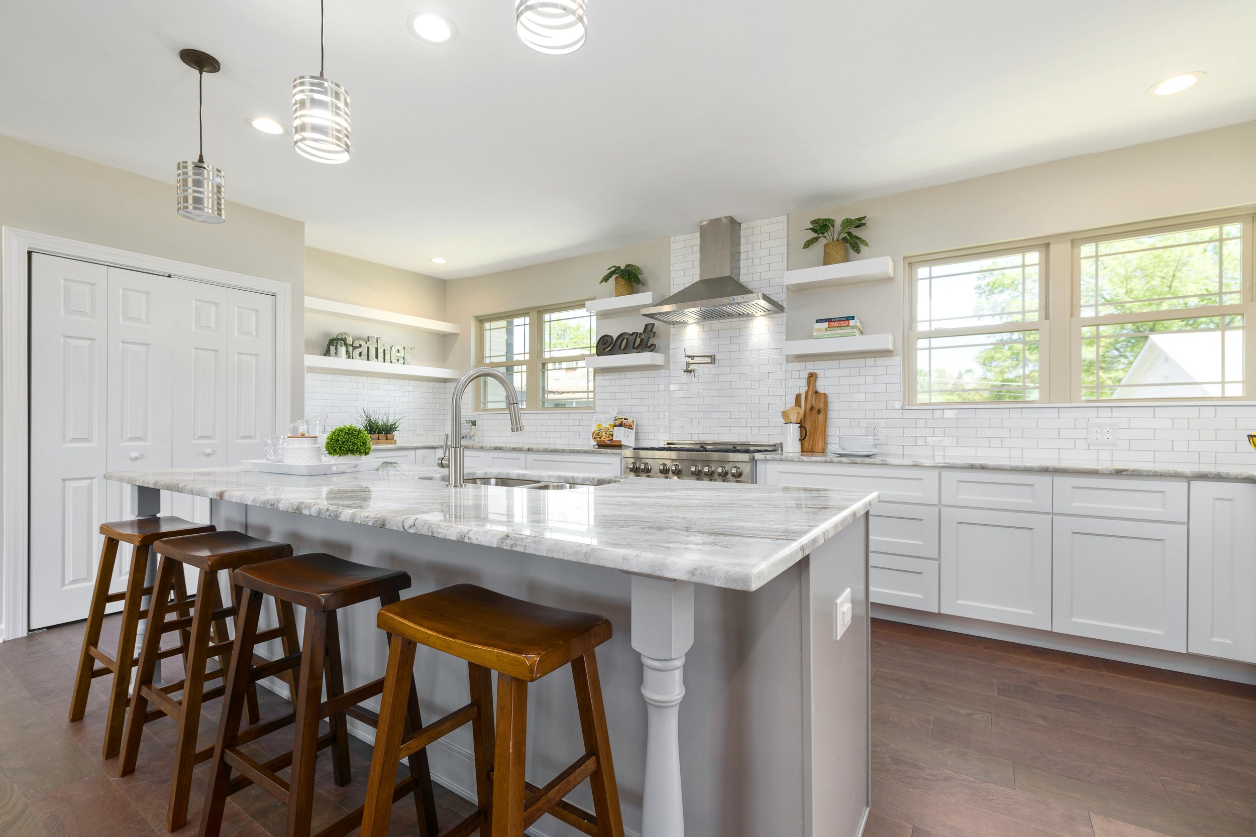 Modern white kitchen with marble island, three wooden stools, stainless steel range hood, white cabinets, open shelves with books and plants, and large windows.