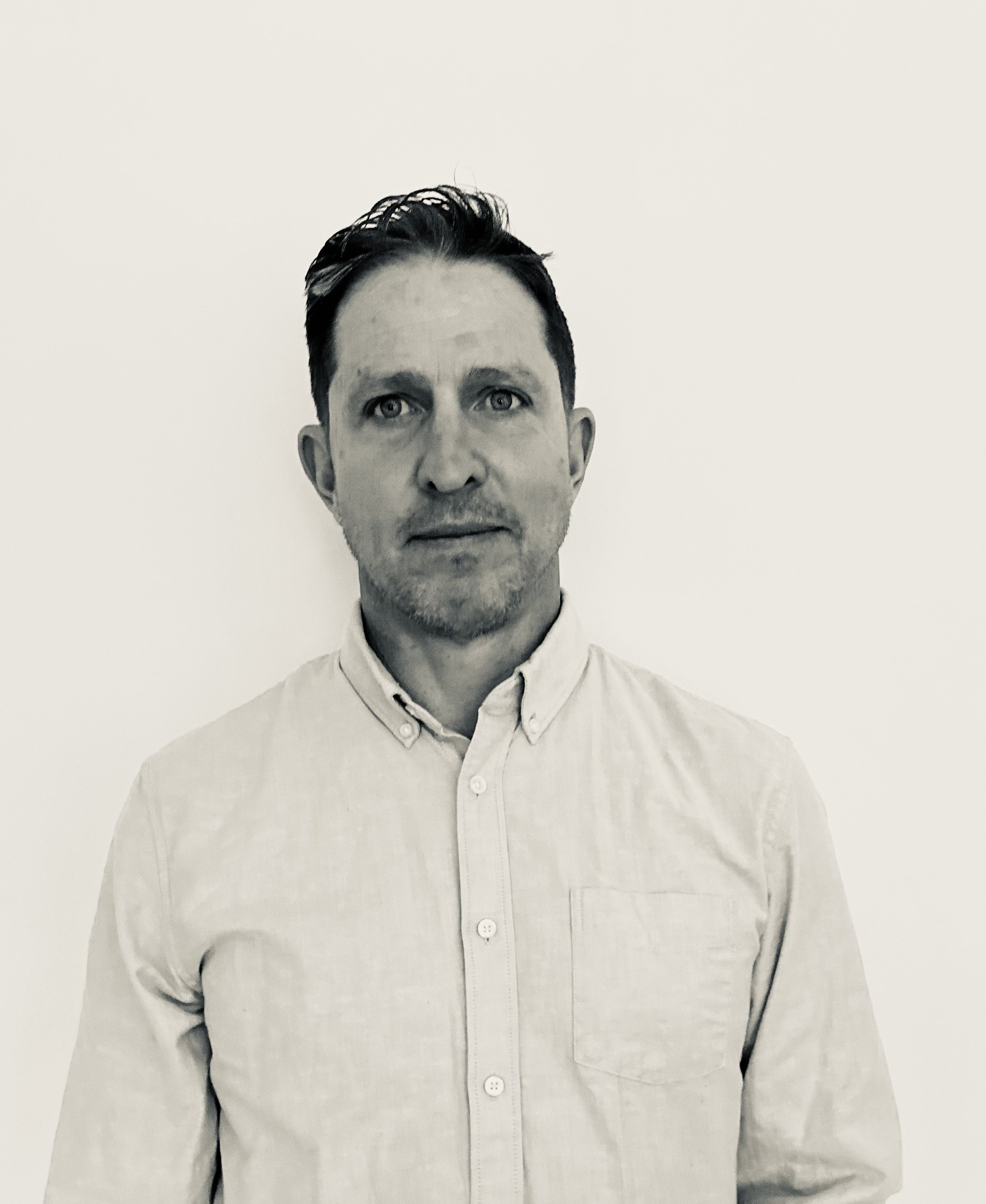 Black and white portrait of a man with dark hair, wearing a light-colored button-up shirt, standing against a plain background.