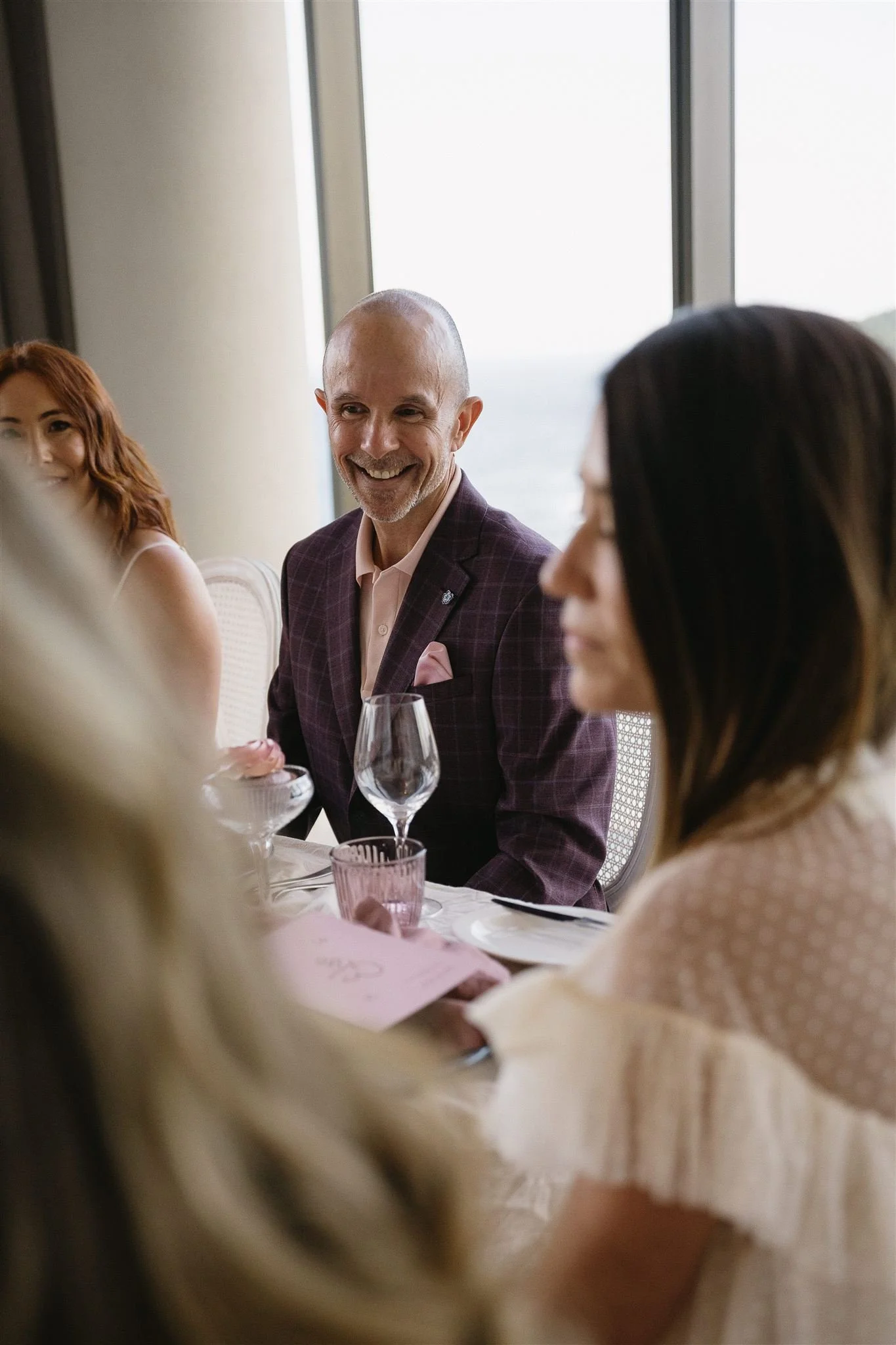 A group of people sitting at a table near a window, engaged in conversation. A man in a suit with a pink shirt and pocket square is smiling at the camera.