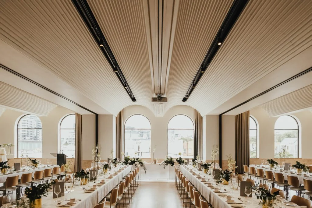 Elegant banquet hall with long tables decorated with white tablecloths, floral centerpieces, and chairs, large arched windows, and high ceiling with wooden panels.