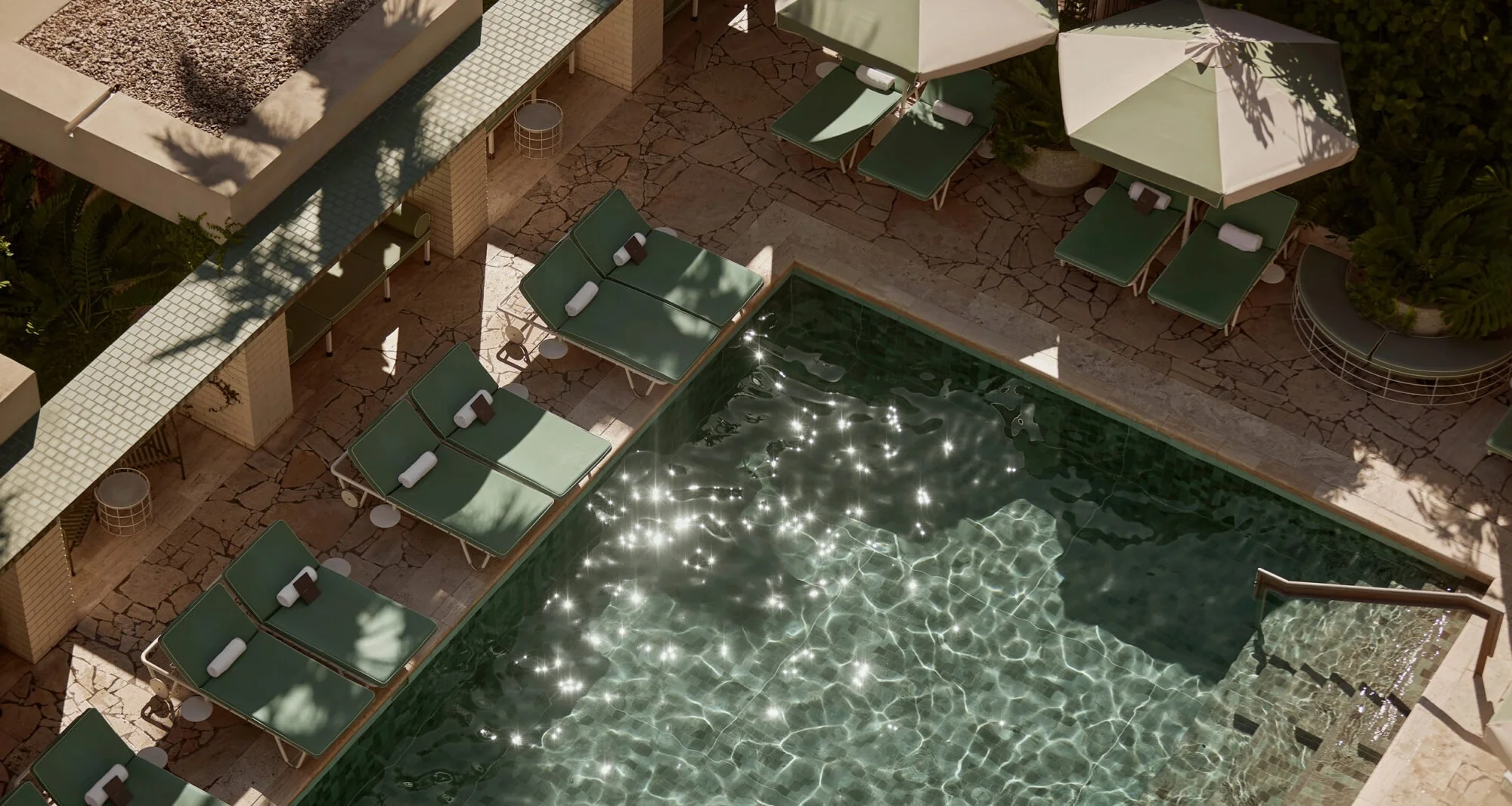 A rooftop swimming pool area with several green lounge chairs and umbrellas, some with white towels on them, and tiny tables on a stone-paved deck, with sunlight sparkling on the water surface.