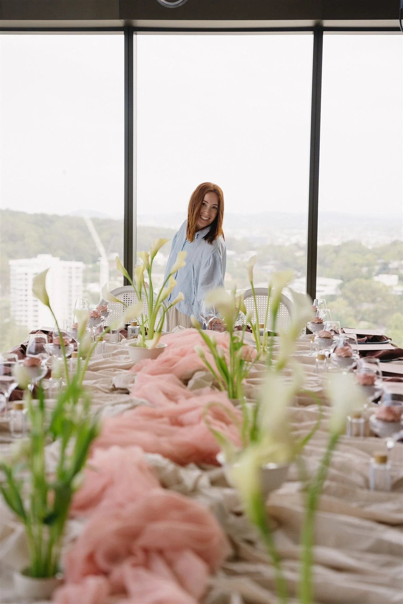 A woman with red hair smiling at a decorated table with calla lilies and pink fabric, in front of large windows with a city view.