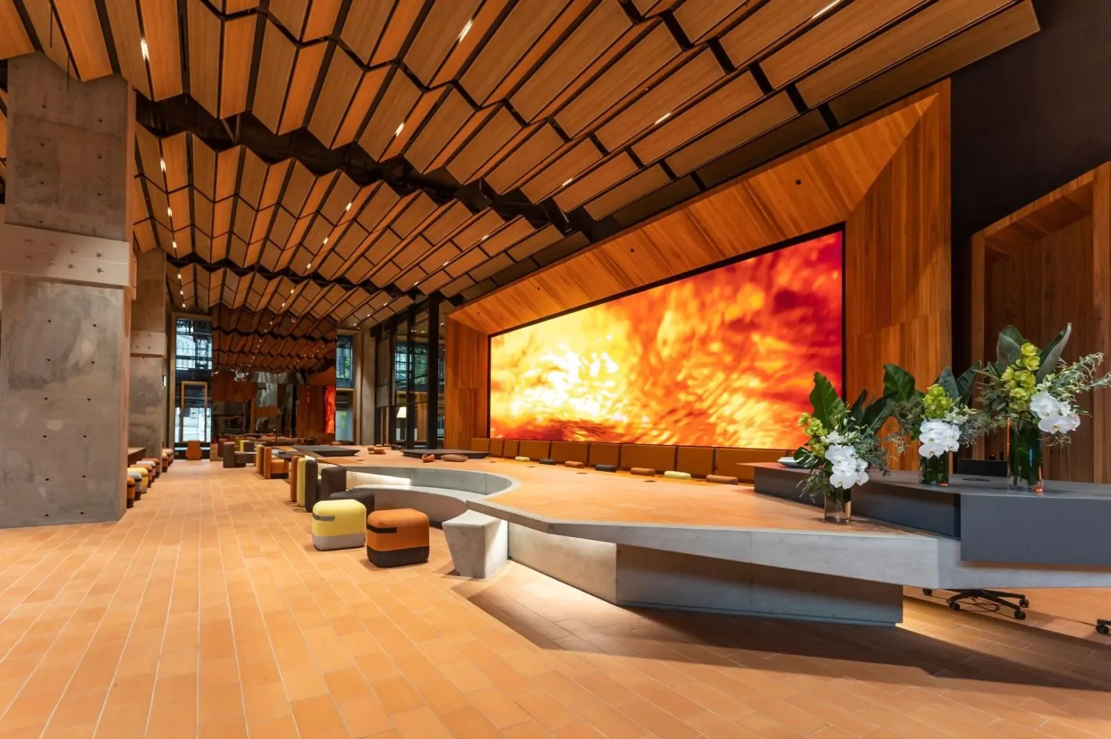 Modern interior with a large digital screen displaying a fiery orange and yellow abstract image, wooden ceilings, and a long reception counter decorated with white flowers in vases.