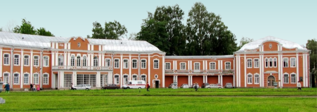 Large historic building with an orange and white facade, multiple windows, and a central balcony, surrounded by a green lawn with people and parked cars in front.