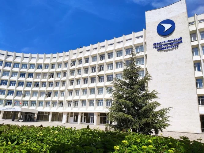 Exterior of a large white academic building with a curved facade, featuring a blue and white emblem and sign reading "Sevastopol State University" in Russian, with green bushes and a pine tree in front under a blue sky.