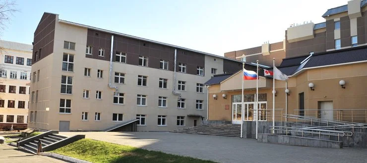 A modern multi-story building with beige and brown exteriors, featuring windows and ramp access, with three flags (one resembling the Texas state flag) flying in front, under a clear blue sky.