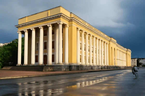 A large, yellow building with tall columns and a classical architectural style, situated at the corner of a wet street with a person walking nearby, under dark cloudy skies.