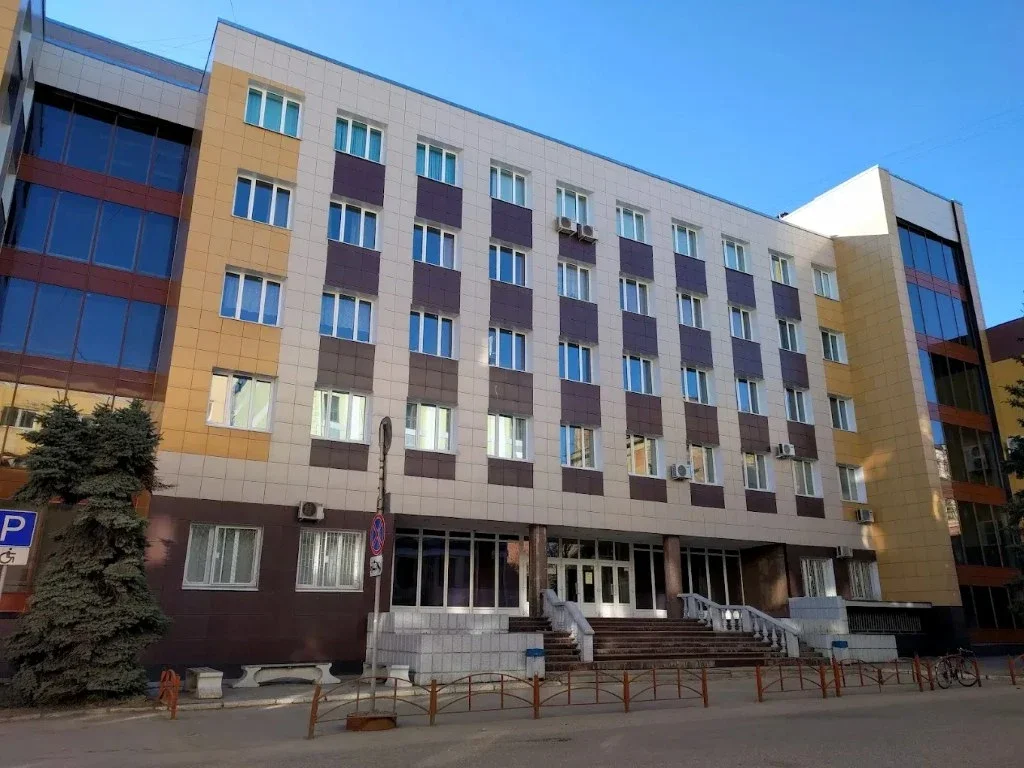 A multi-story building with a beige and brown tiled facade, multiple windows, and air conditioning units. The entrance has a stairway and glass doors. There are trees, a bench, and a bicycle outside.