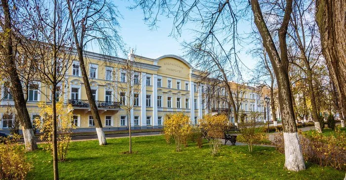 A yellow three-story building with white accents and arched windows, surrounded by trees and a grassy park with benches in the foreground.