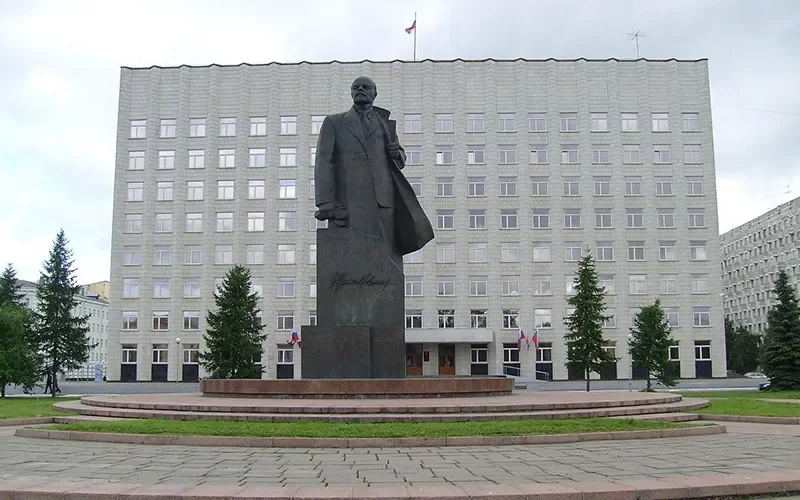 A large statue of a man stands in front of a government building, with trees and flags around it.