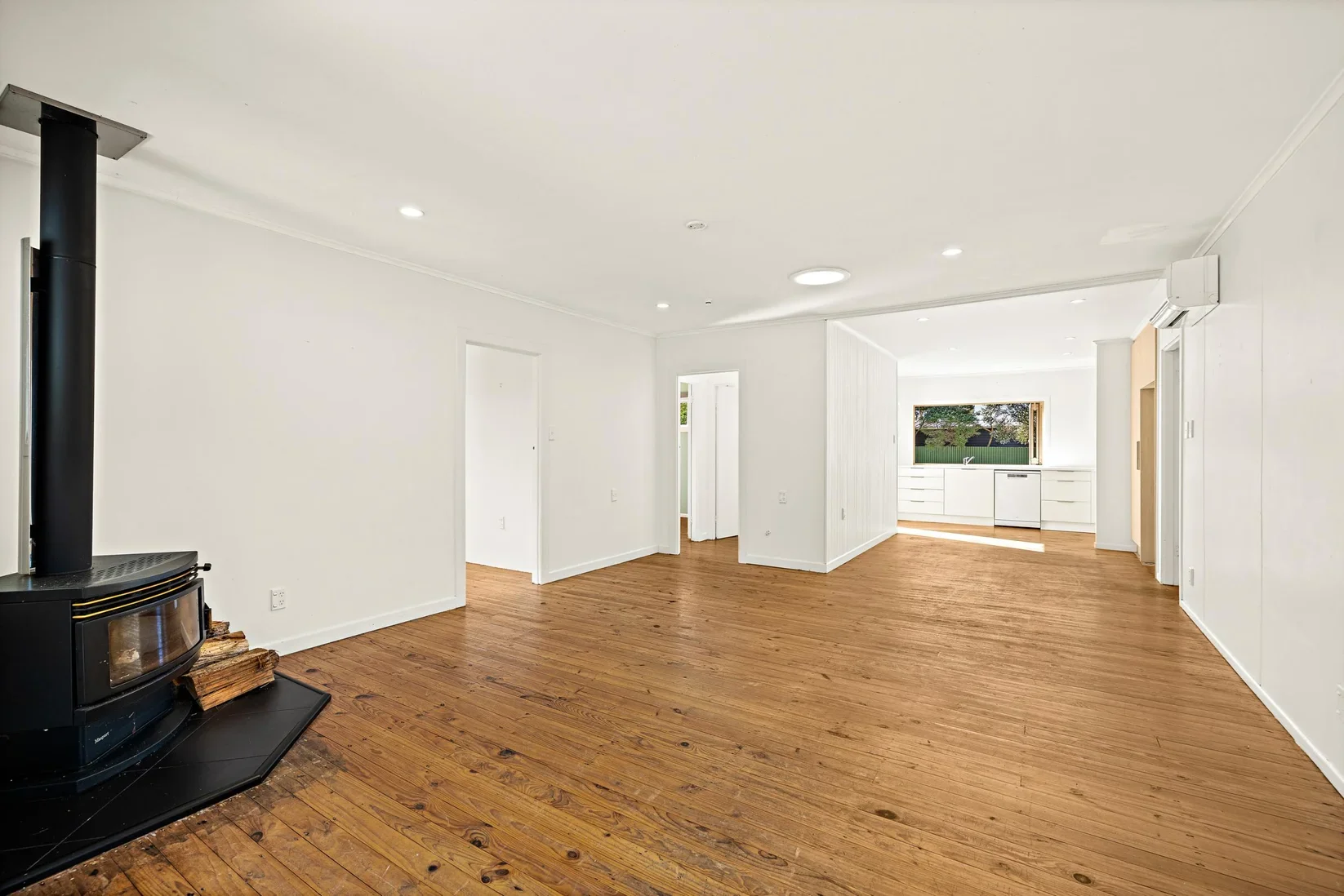 Empty living room with wooden floors, white walls, a black wood stove, and a partial view of the kitchen with a window showing trees outside.