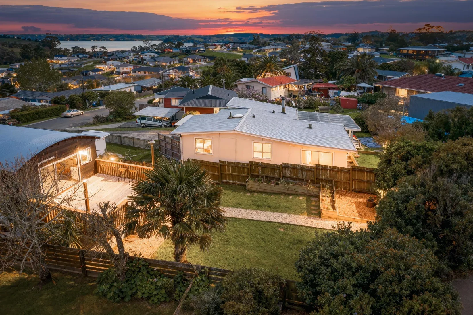 A neighborhood at dusk with houses, trees, and a lake in the background. The house in the foreground has a lit backyard patio and a palm tree.