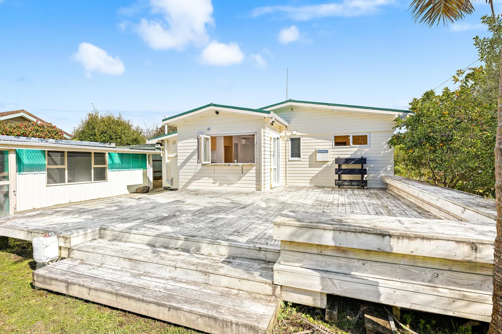Backyard in Point Wells with a spacious wooden deck, a white house with a corner window, opened shutters, and a small porch. There are trees and shrubs around, under a blue sky with some clouds.