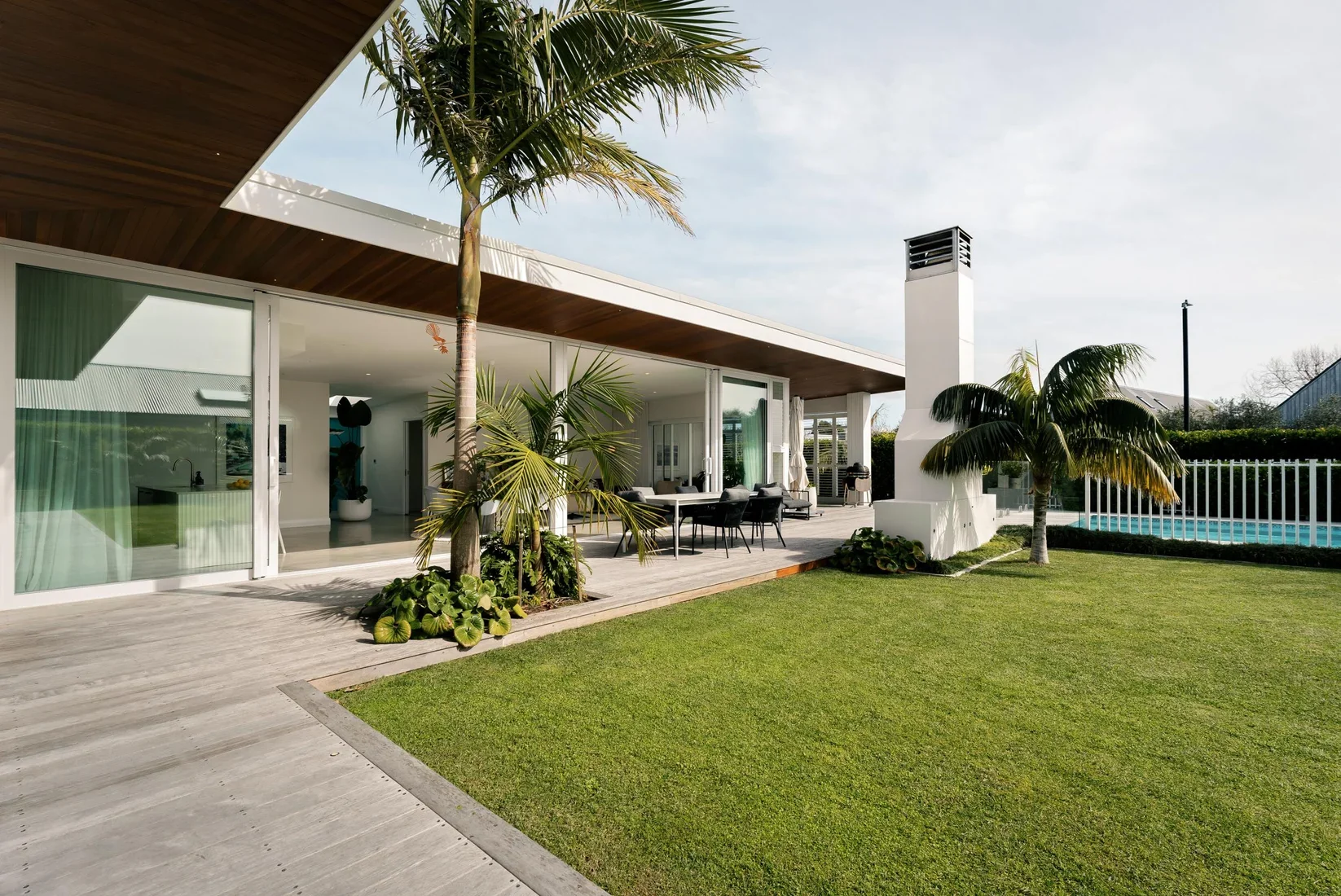 Modern house with large glass doors, outdoor patio with black chairs and table, green lawn, palm trees, and a swimming pool enclosed by a white fence.