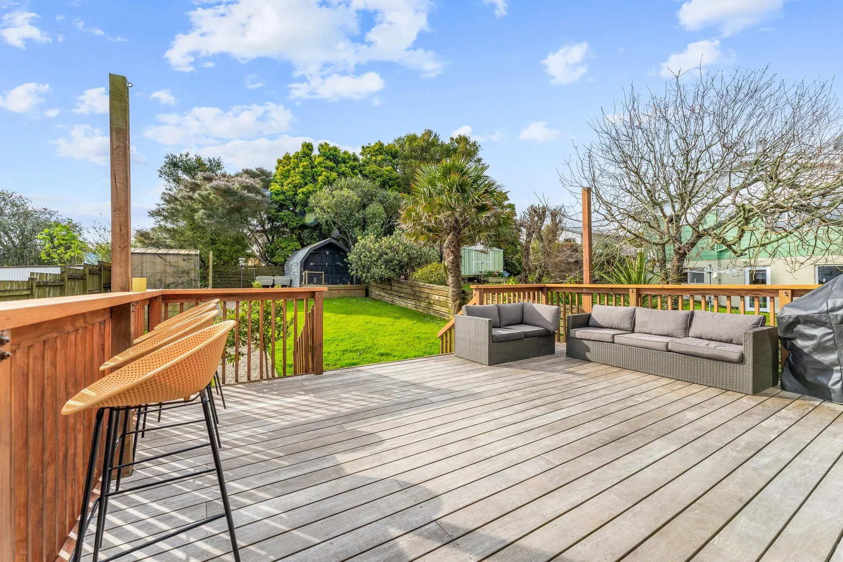 Wooden deck with outdoor seating, including two gray sofas and a black sofa, overlooking a green backyard with trees and a shed, under a partly cloudy sky.