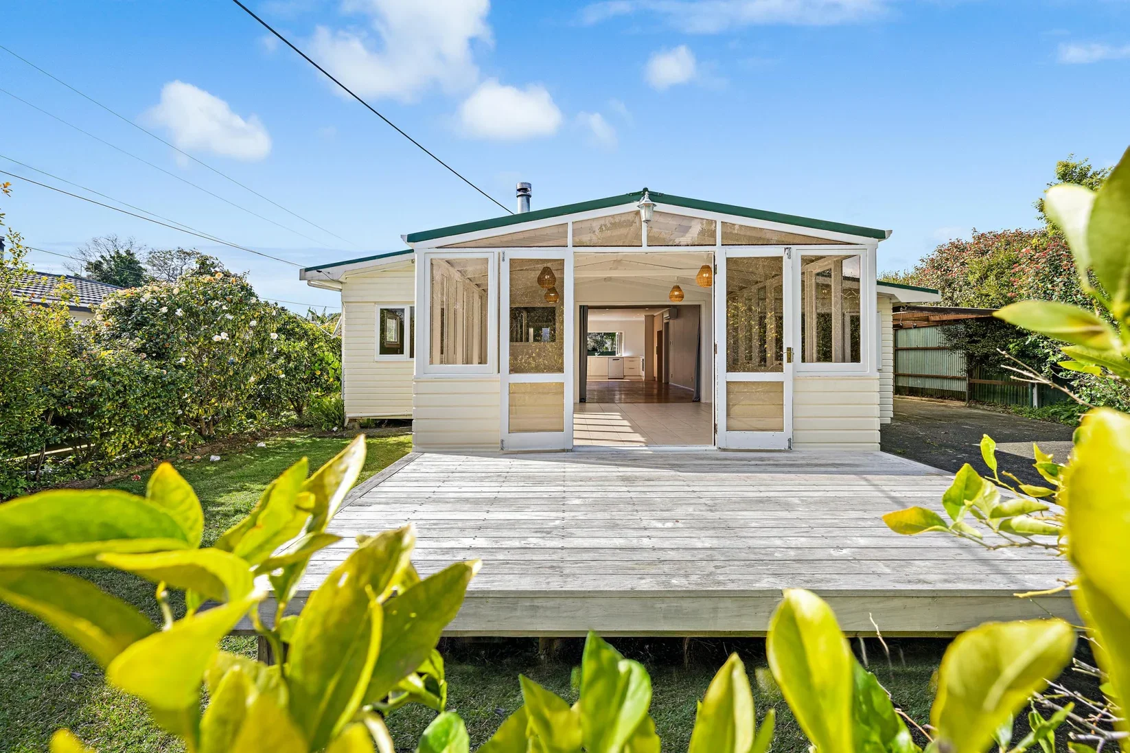 View of a house with a glass sunroom opening onto a small wooden deck, surrounded by green plants and shrubs, with a blue sky and fluffy clouds overhead.