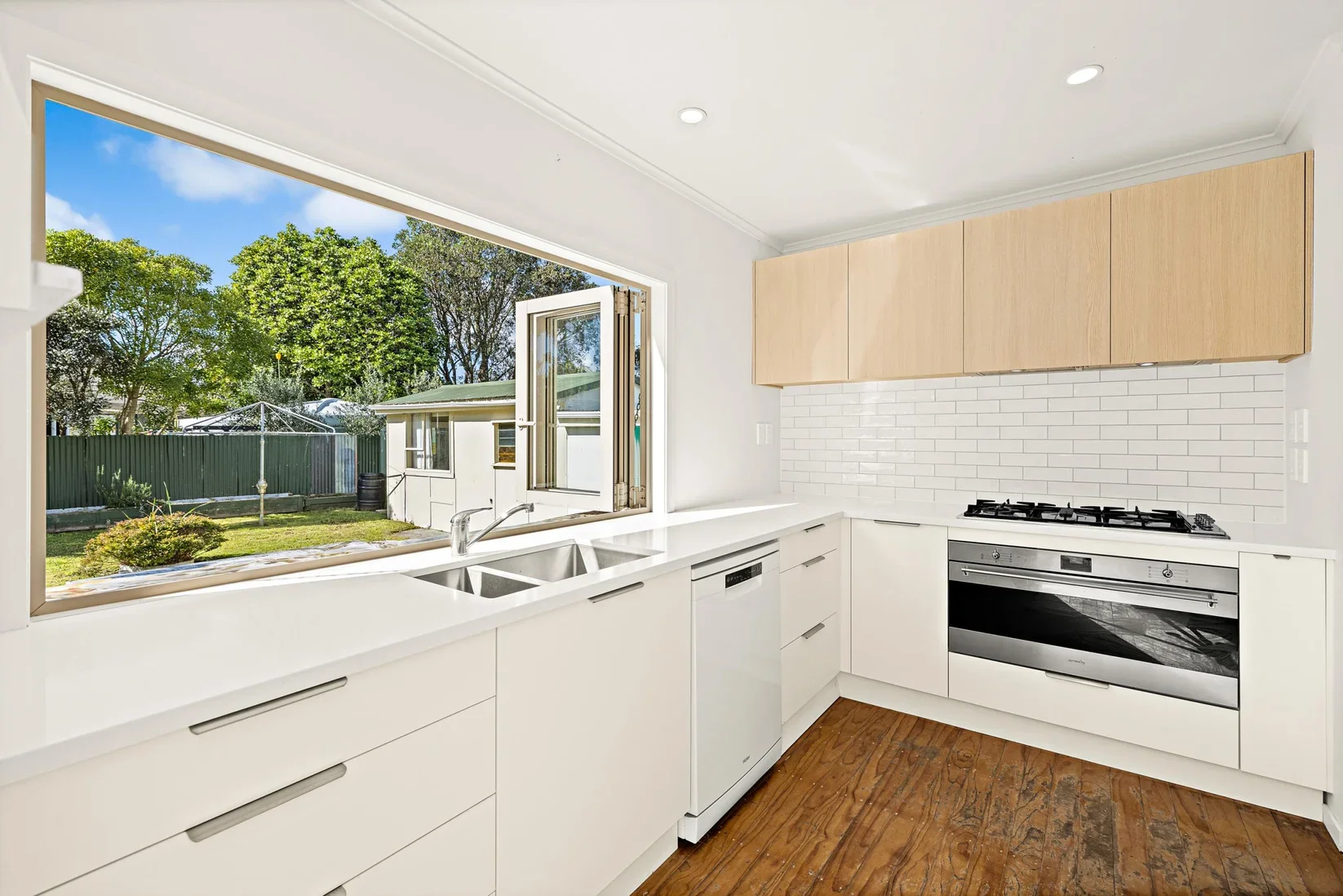 Modern kitchen with white cabinets, stainless steel oven, and a large window overlooking a backyard with trees and a clothesline.